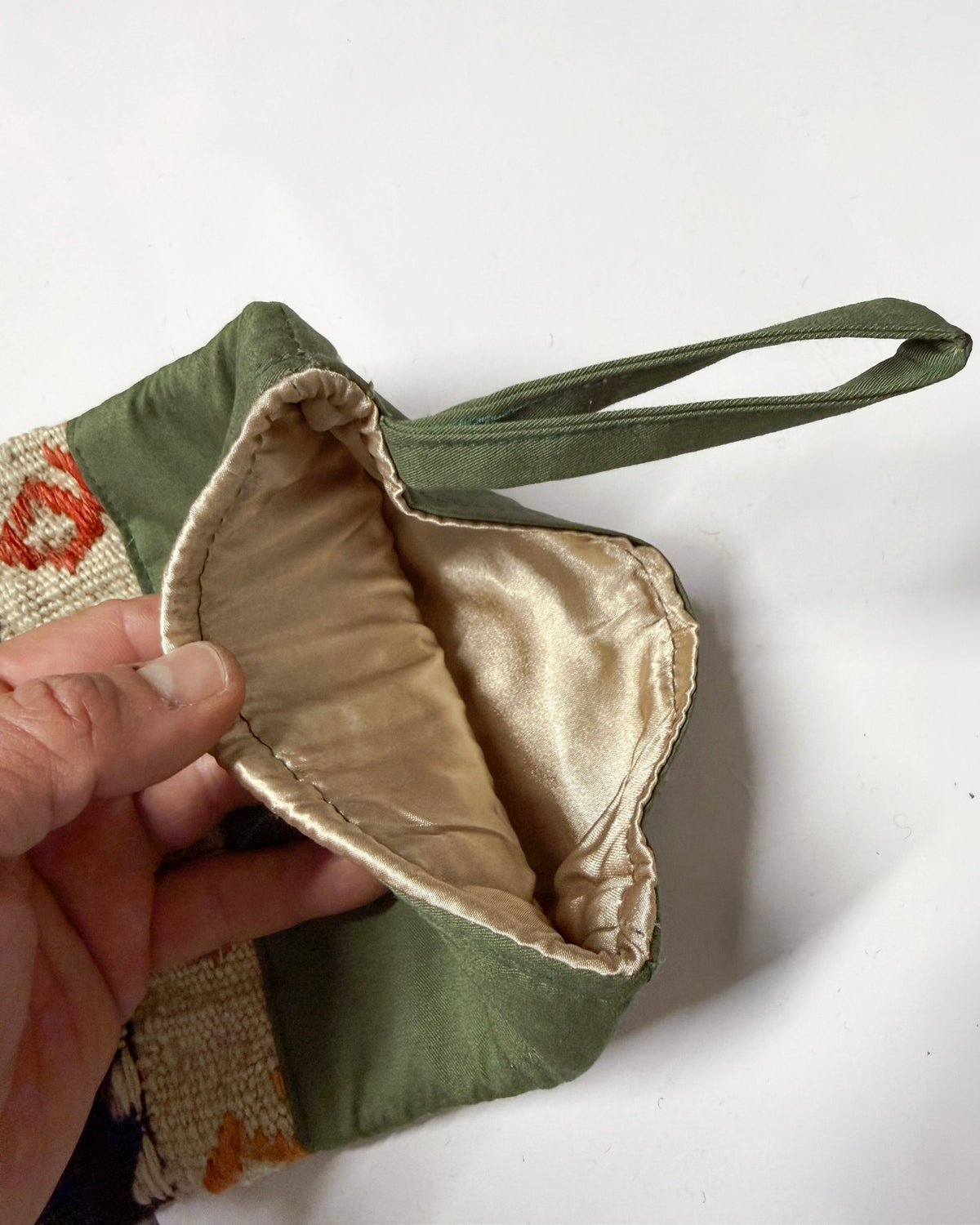 A close-up of a hand holding open a small fabric bag, showing an olive green exterior and a shiny beige satin interior lining. The bag has a matching green handle and a patterned textile edge visible on the left.