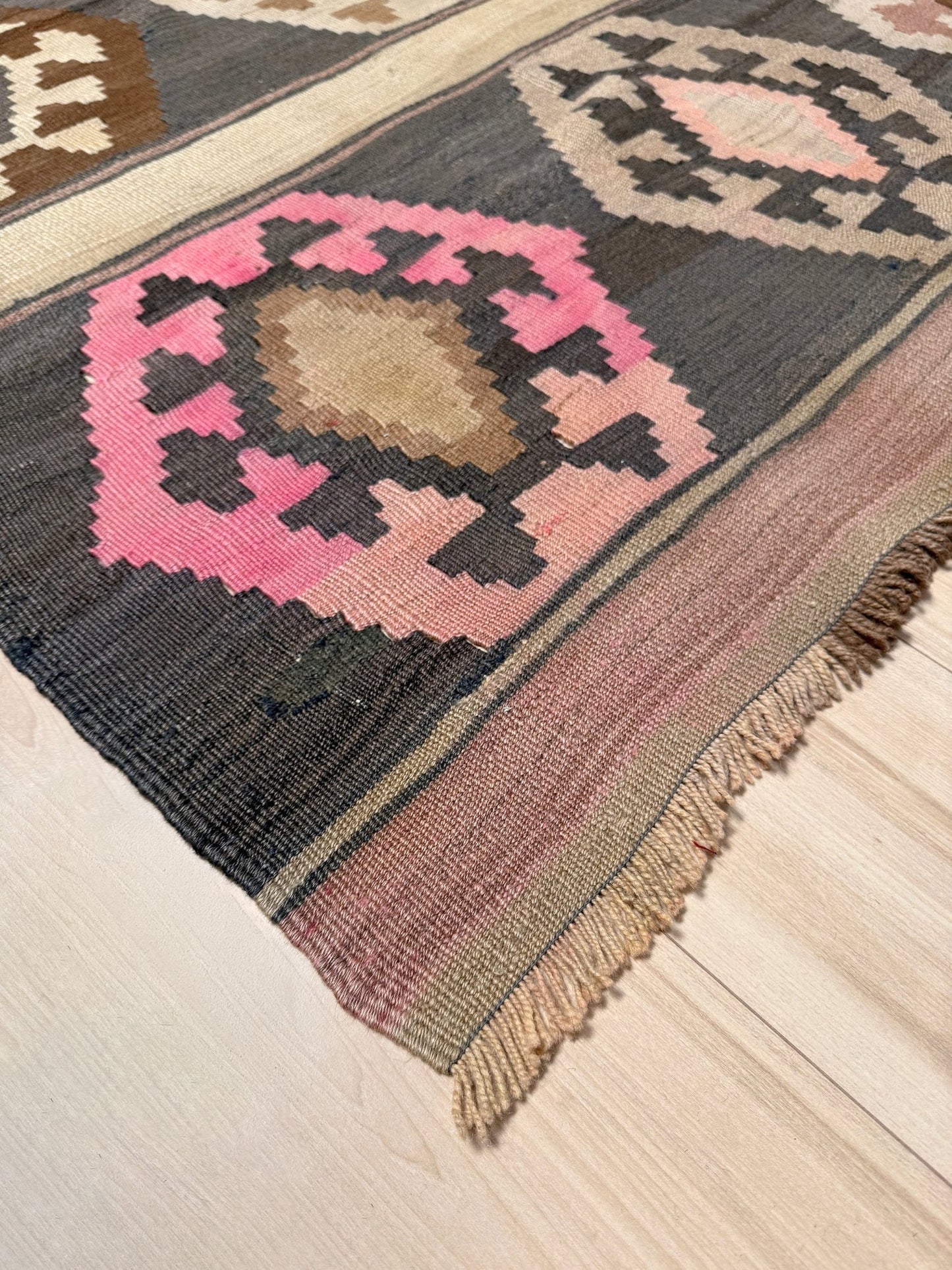 A close-up of a woven area rug with a geometric pattern in pink, beige, brown, and black, laid out on a light wood floor. Handmade wool 6x9 rug from Indigo Rugs Burlingame Bay Area.