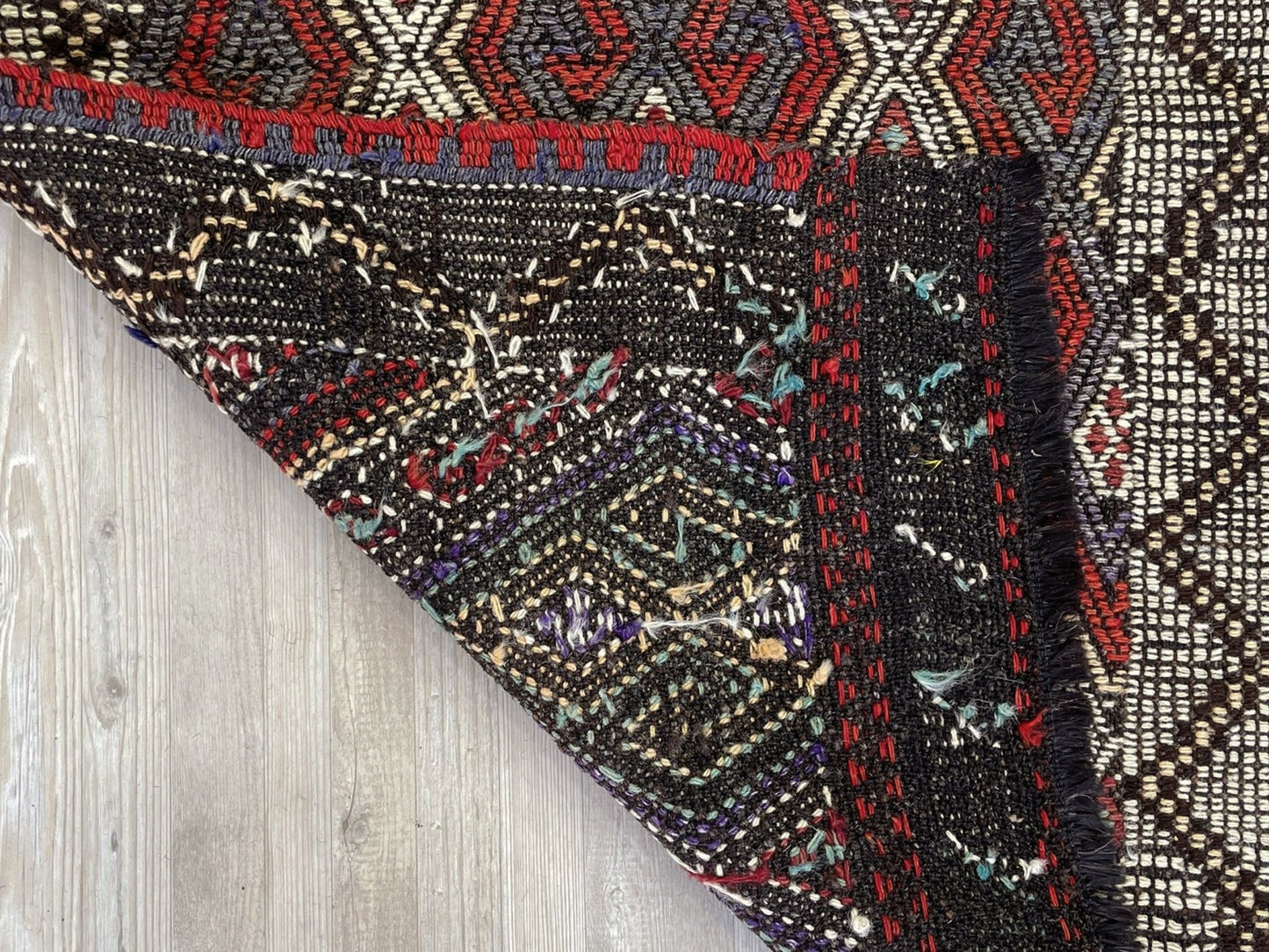 Close-up of a patterned fabric with intricate designs on a wooden surface. Small vintage Turkish cicim kilim rug, 2x4, with raised tribal embroidery in rust red, ivory, olive, and charcoal on a goat hair foundation.