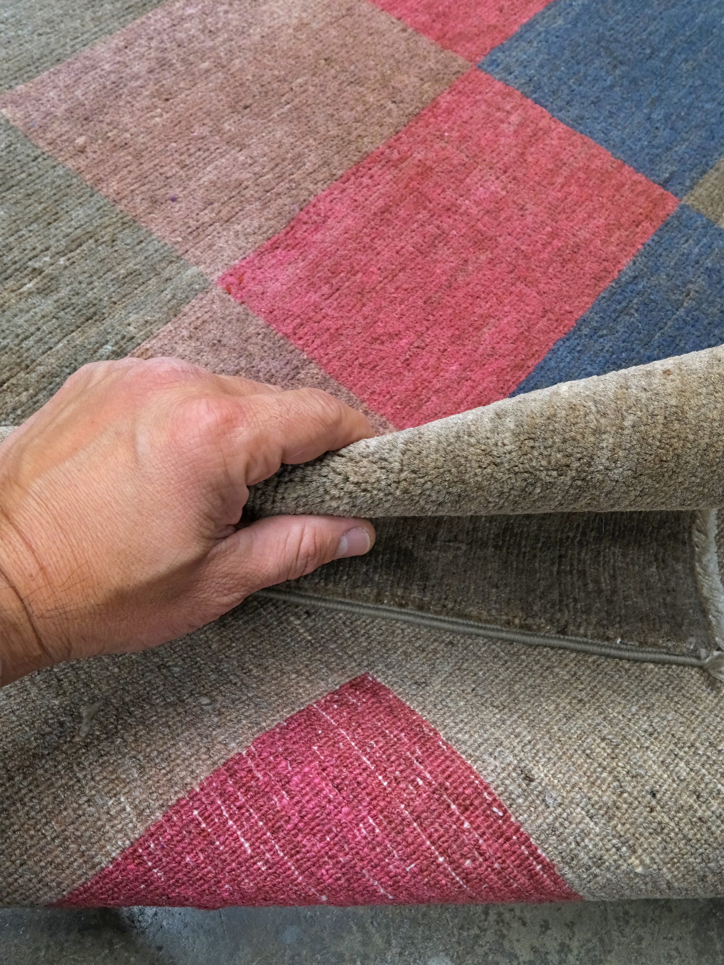 A close-up of a hand lifting the corner of a woven rug to show its texture and backing. The rug features a diamond pattern in muted beige, dusty pink, blue, and olive green.