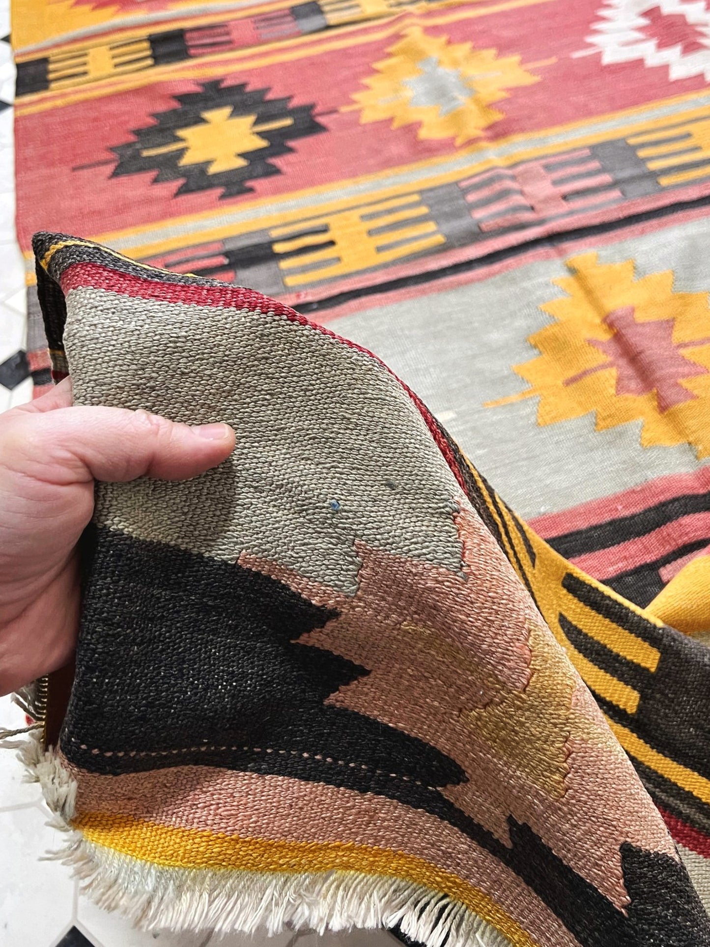 Hand holding a piece of fabric with geometric patterns in front of a colorful patterned rug. Vintage Afyon Turkish kilim runner, 4x9, with striped tribal design in muted brick red, dusty rose, bright marigold, and charcoal tones.