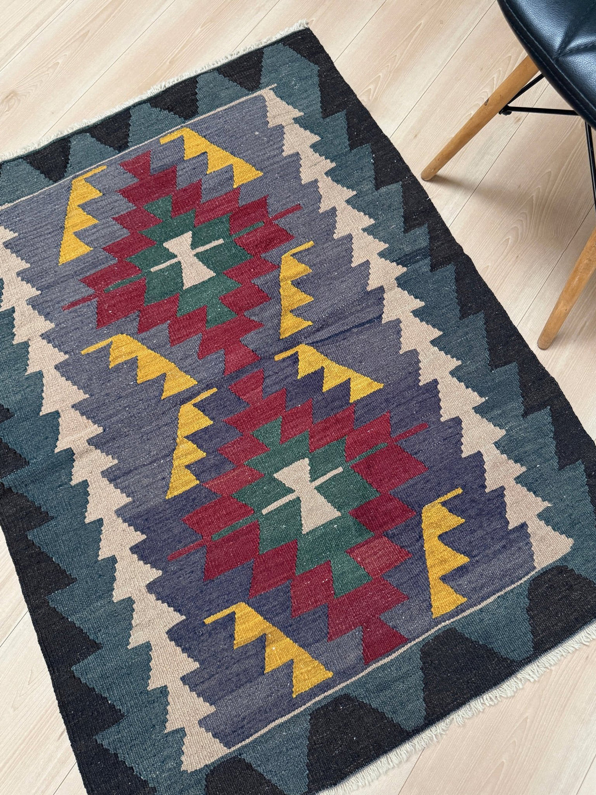 A rectangular woven area rug with a Southwestern geometric pattern, shown on a light wood floor next to a black chair.