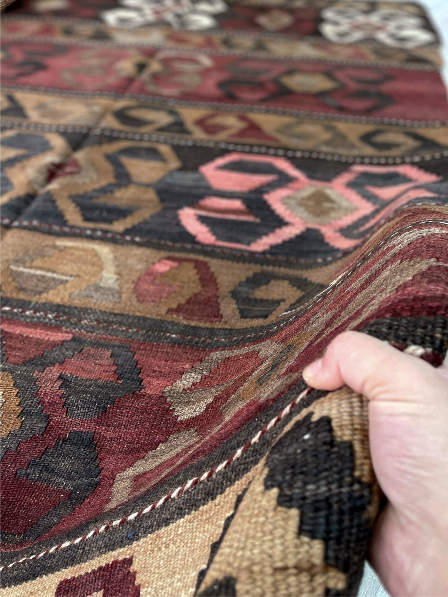 Close-up of a hand holding a patterned rug with intricate designs. Vintage Eastern Turkish Kars kilim rug with horizontal tribal bands, hooked diamond motifs, and geometric symbols in madder red, walnut brown, 5x7.