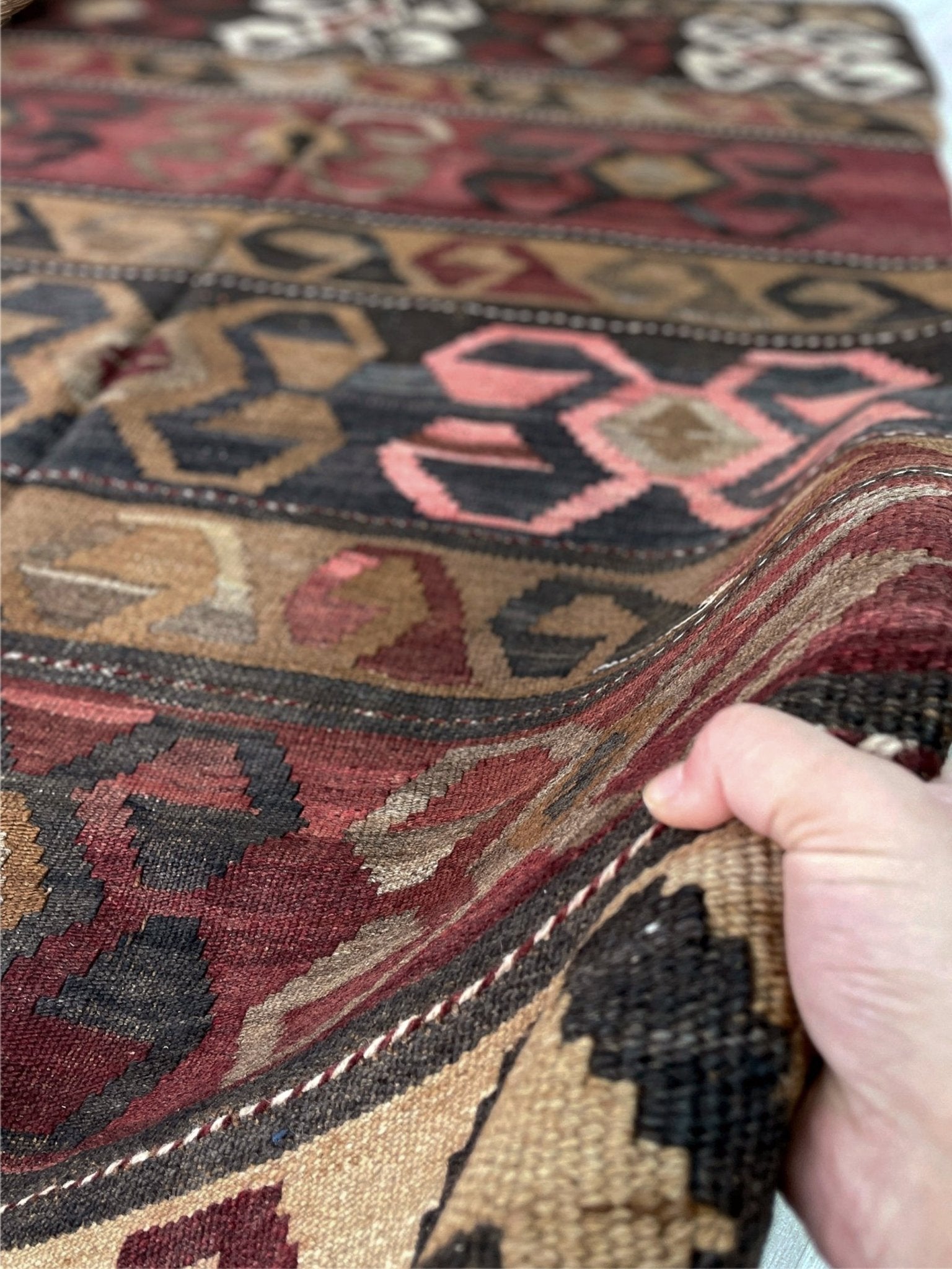 Close-up of a hand holding a patterned rug with intricate designs. Vintage Eastern Turkish Kars kilim rug with horizontal tribal bands, hooked diamond motifs, and geometric symbols in madder red, walnut brown, 5x7.