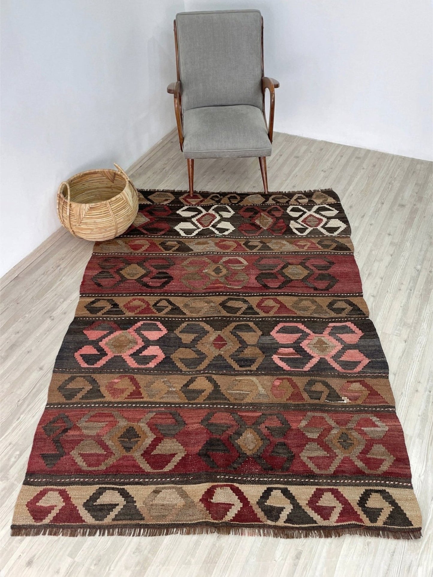 Woven rug with geometric pattern on a wooden floor, with a chair and basket in the background. Vintage Eastern Turkish Kars kilim rug with horizontal tribal bands, hooked diamond motifs, and geometric symbols in madder red, walnut brown, 5x7.