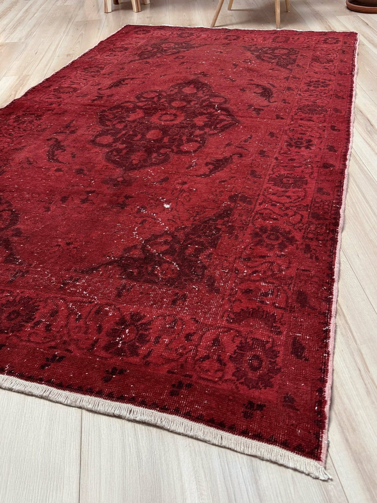 A rectangular red area rug with a distressed traditional floral pattern and cream fringe, laid out on a light-colored wooden floor. Moody red overdyed Turkish rug with distressed vintage pattern, deep burgundy red field, tonal medallion and border design, handmade wool accent rug 5x7 from Indigo Rugs Burlingame Bay Area
