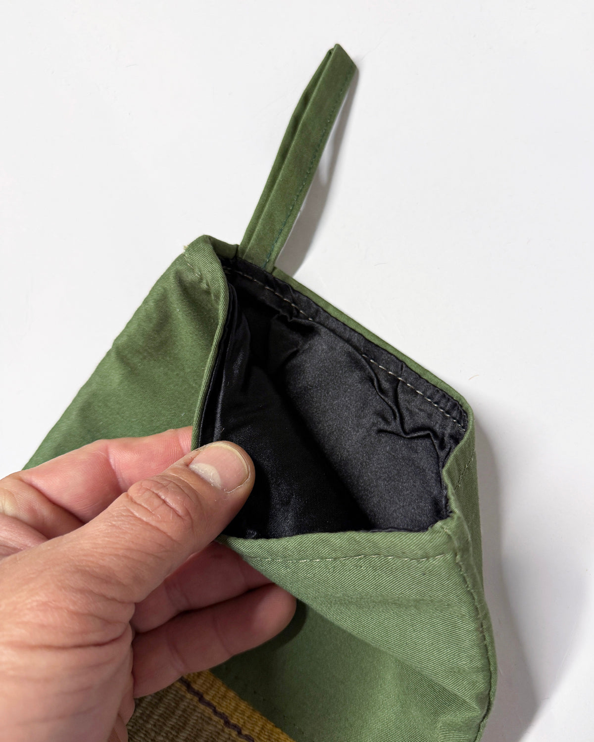 A close-up of a hand holding open an olive green fabric pouch or small bag, showing a black interior lining against a plain white background.