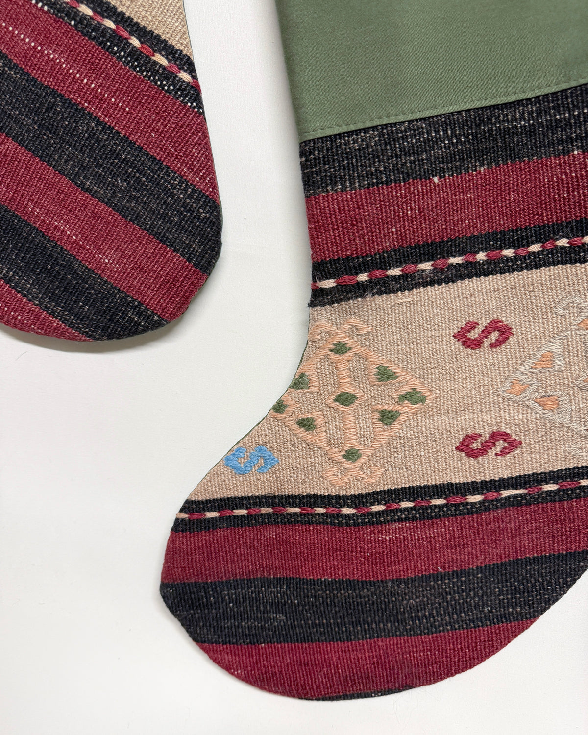 A close-up of a handwoven Christmas stocking with a Southwestern design, featuring stripes of burgundy, black, and beige, with a green top panel and embroidered geometric motifs.