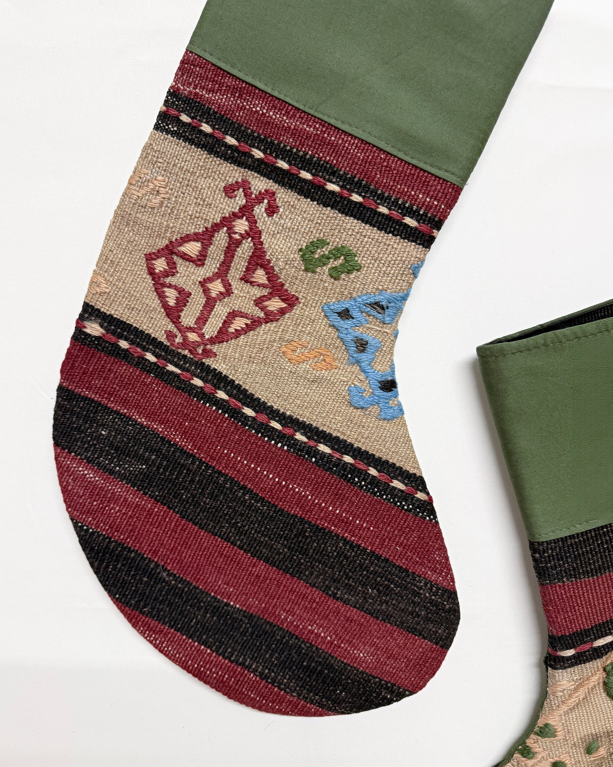 A close-up of a patterned Christmas stocking Turkish Wool Kilim, featuring an olive green cuff, horizontal stripes of black and burgundy, and festive embroidered motifs including a red candy cane and a blue ornament.