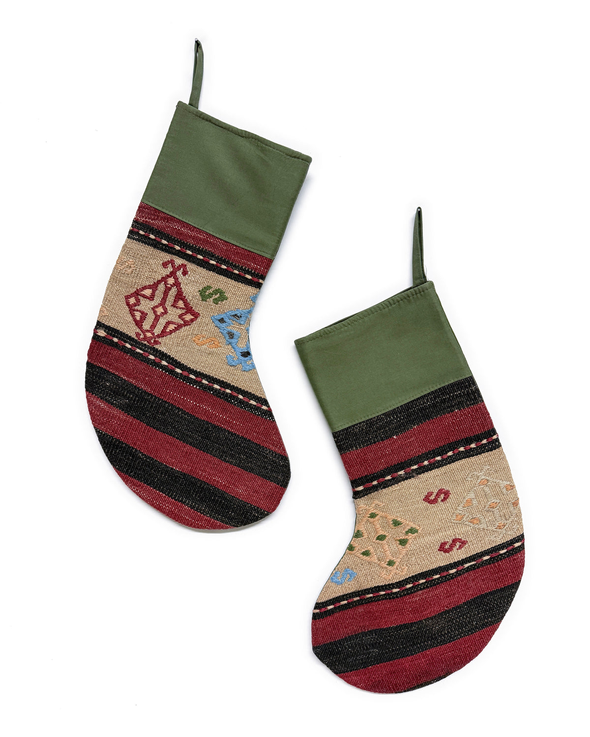 A pair of Kilim Christmas stockings with olive green cuffs, featuring horizontal stripes in red, black, and beige, with small embroidered holiday motifs.