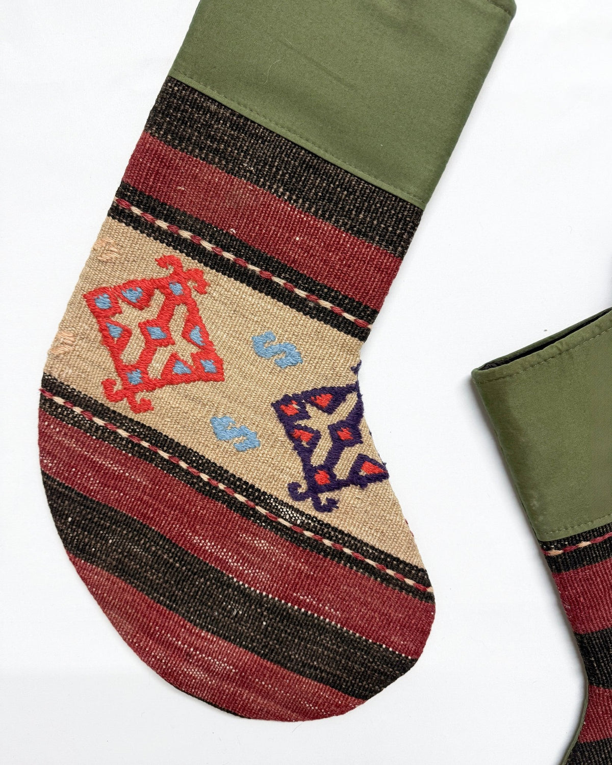 A close-up of a woven Christmas stocking with an olive green cuff, featuring horizontal stripes in red, black, and beige, with embroidered red and blue geometric motifs on a light tan section.