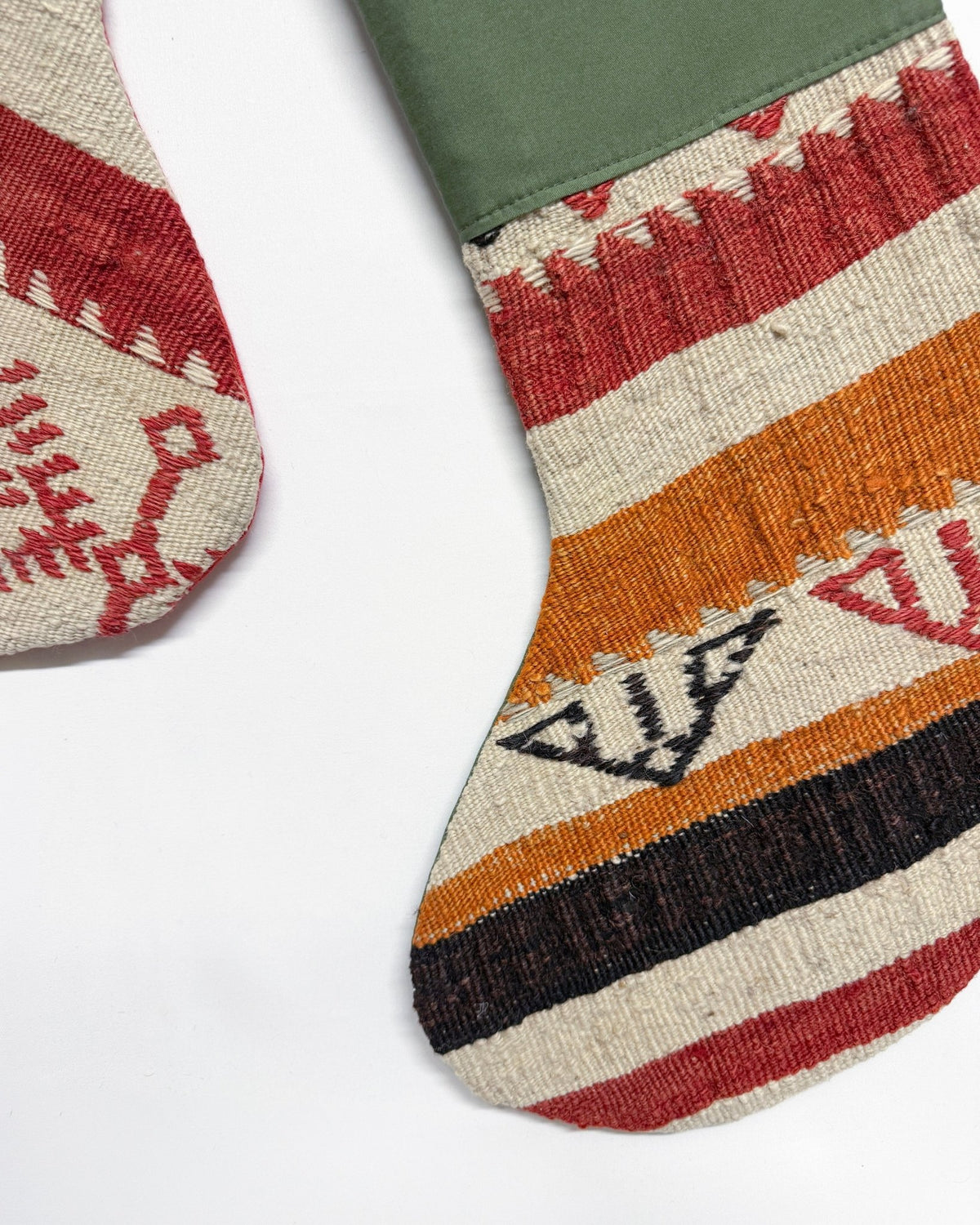 A close-up of a handwoven textile stocking with horizontal stripes in red, orange, black, and cream, featuring geometric patterns and an olive green cuff, shown on a white background.