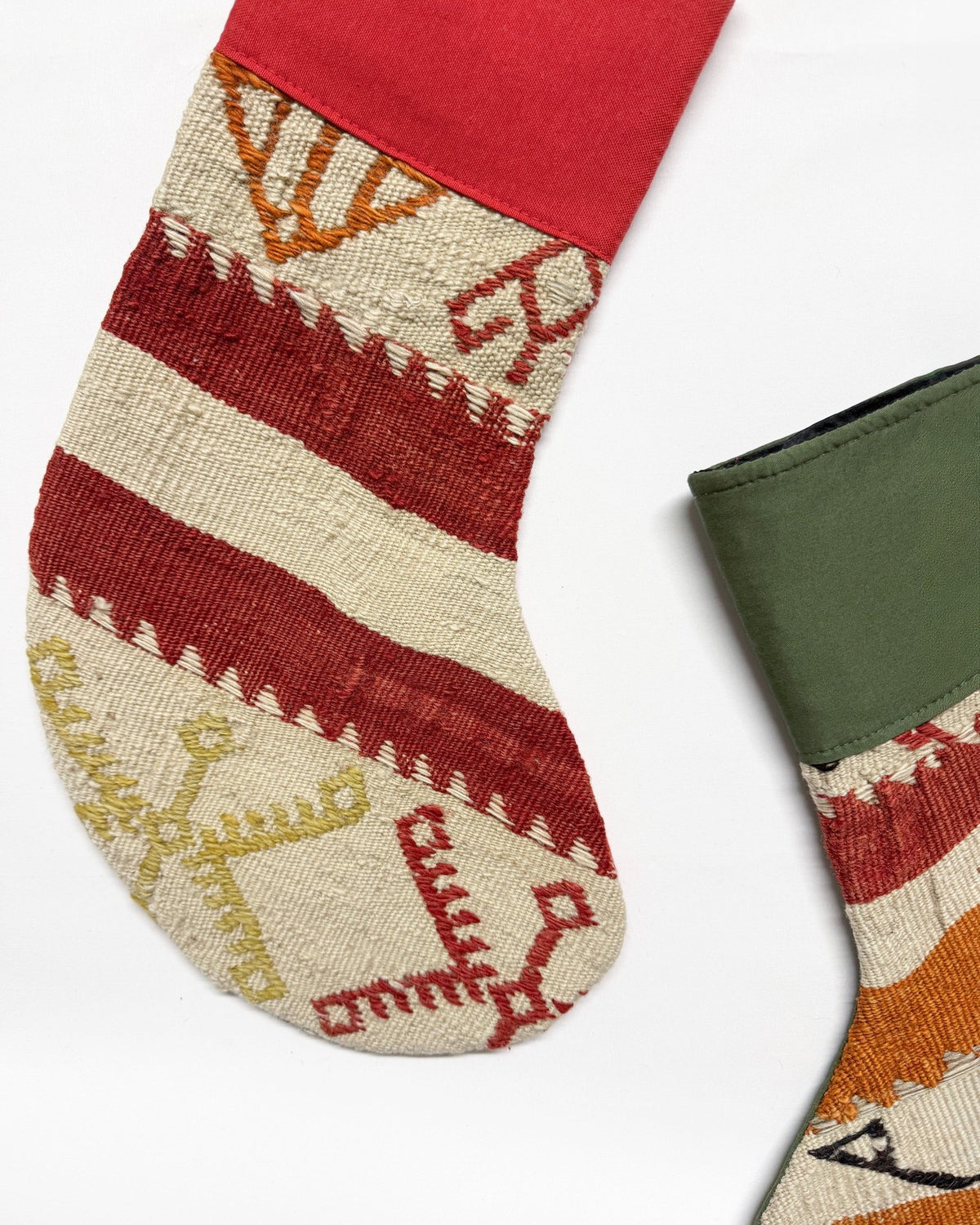 A close-up of a patterned Christmas stocking with horizontal stripes in red, beige, and orange, featuring geometric and animal motifs. A second stocking is partially visible on the right, showing an olive green cuff.