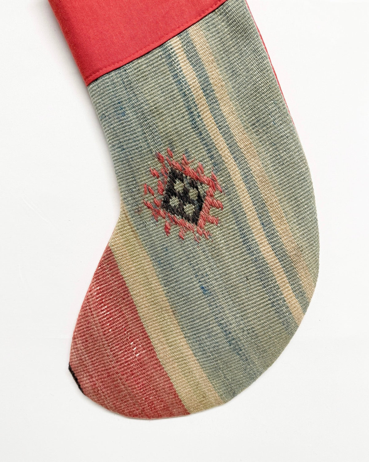 A Kilim handmade Christmas stocking featuring a red cuff and hanging loop, with a body made from striped kilim fabric in sage green, beige, and rust red, accented by a small geometric woven motif.