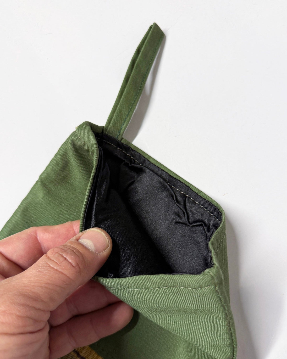 A close-up of a hand holding open an olive green fabric pouch to show its black interior lining, set against a plain white background.