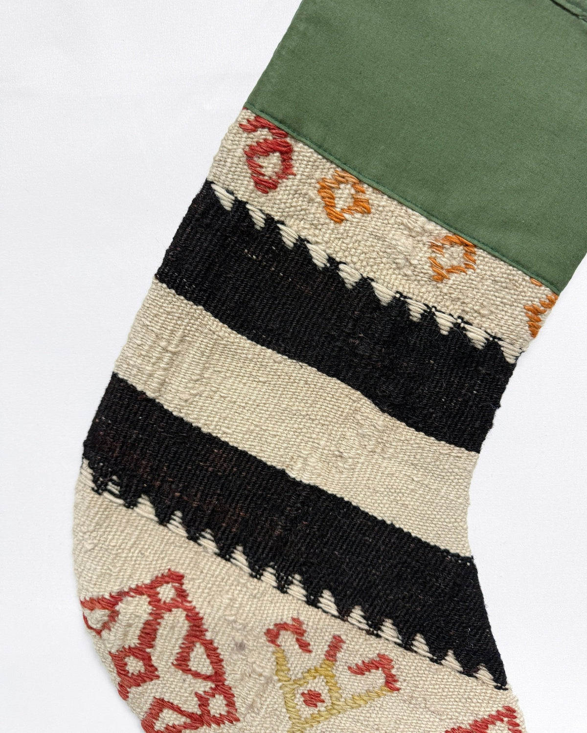 A close-up of a handwoven Christmas stocking with horizontal stripes of black and cream, accented with red and orange geometric patterns, and finished with an olive green cuff.