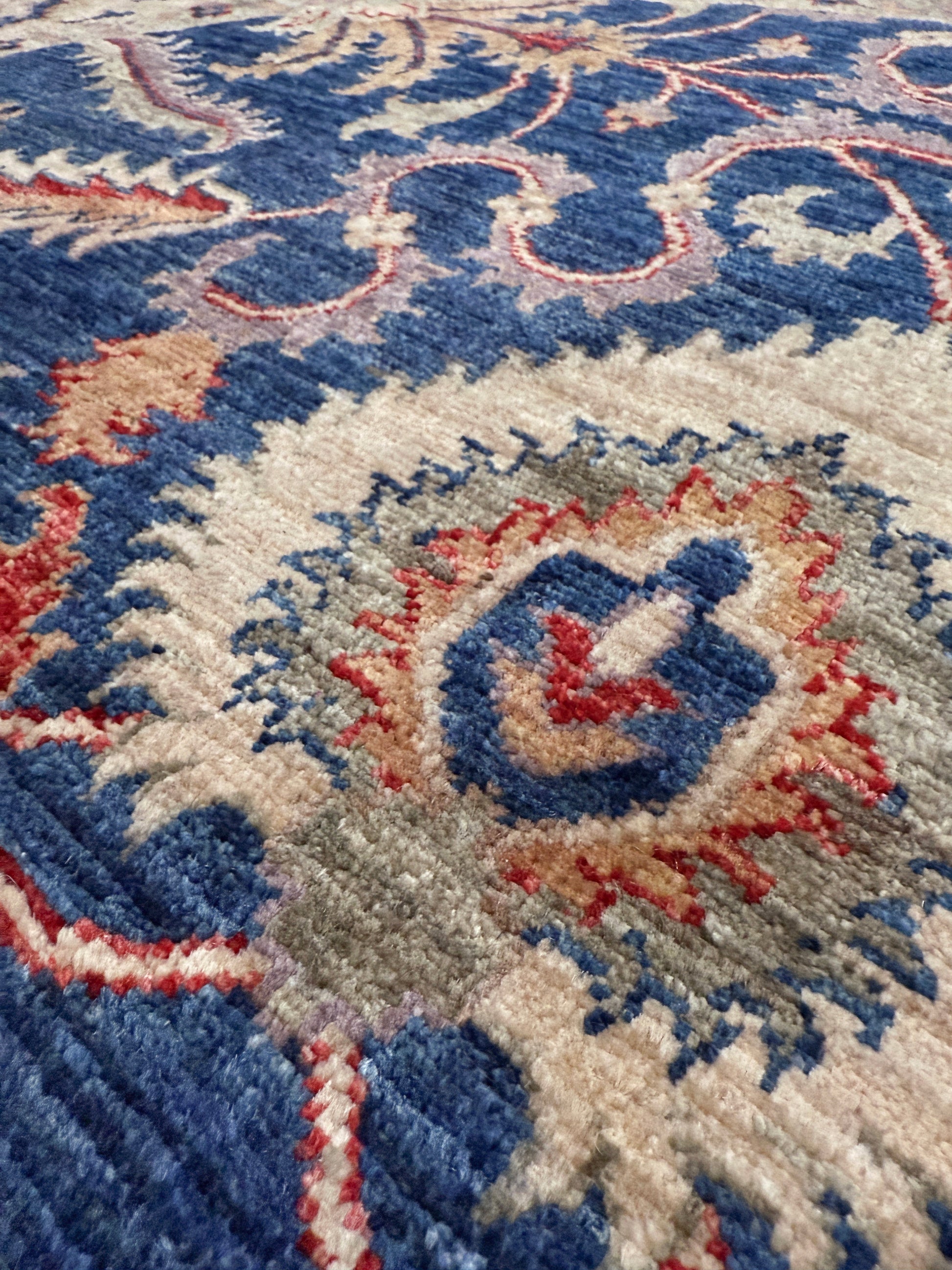 Close-up of a patterned rug with blue, red, and beige colors. Round Oushak rug with blue field, beige border, muted gold, dusty red floral details, handmade wool 6x6 round area rug from Indigo Rugs Burlingame Bay Area.