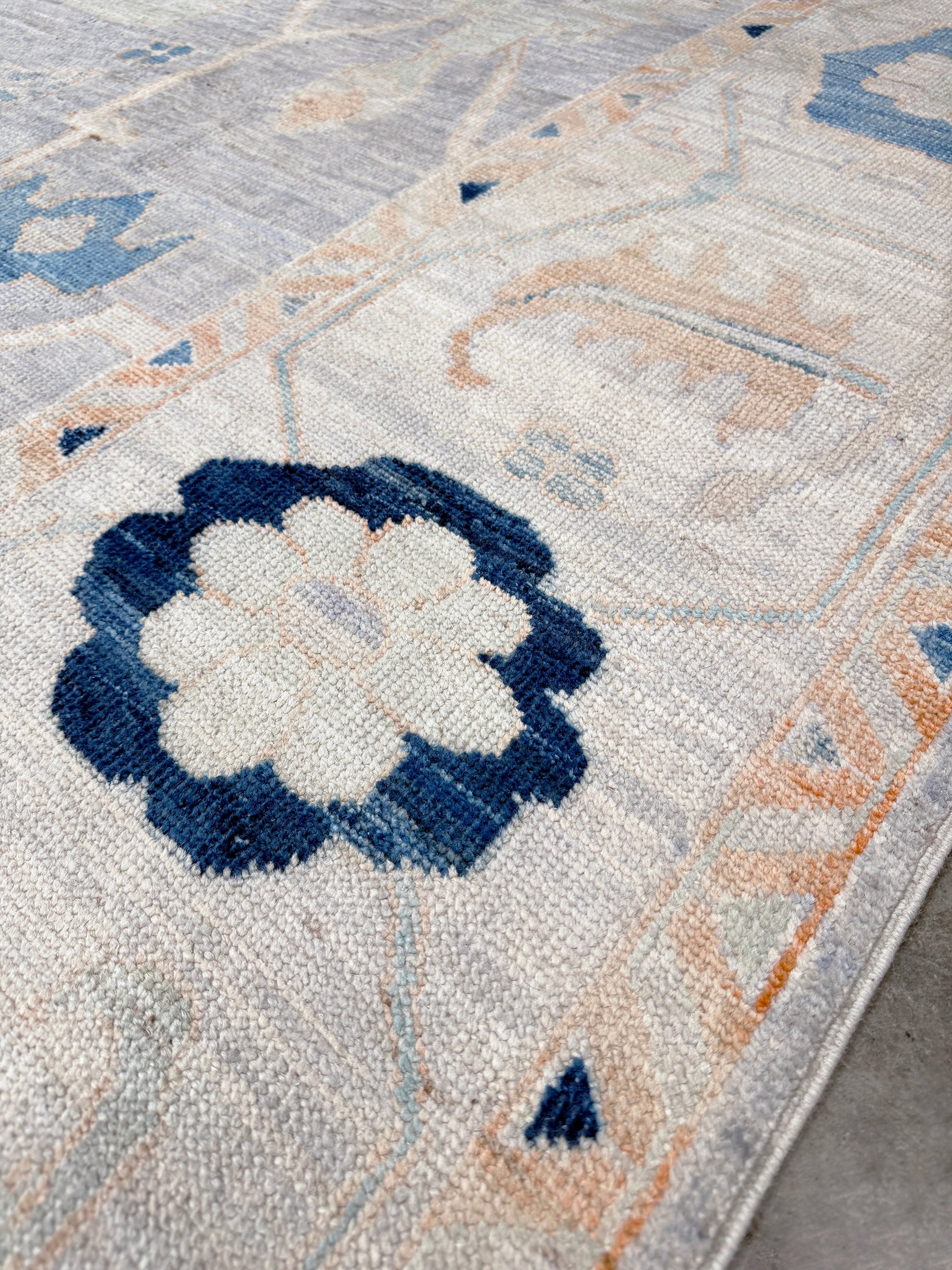 Close-up of a patterned rug with a blue and white floral design. Brand new Turkish Oushak rug with pale blue field, subtle lilac undertones, soft ivory border, faded blue and peach beige accents, handmade wool area rug 9x12 from Indigo Rugs Burlingame Bay Area.