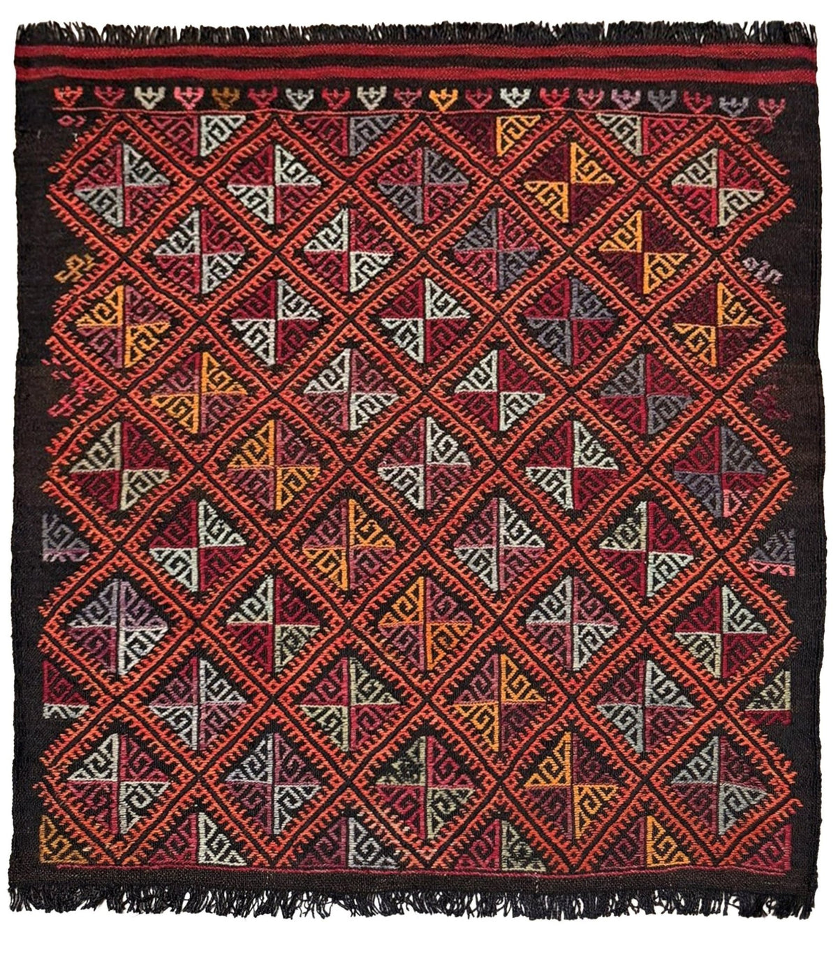 Decorative square turkish kilim rug with geometric pattern on a wooden floor, black chair, and potted plant in the background.