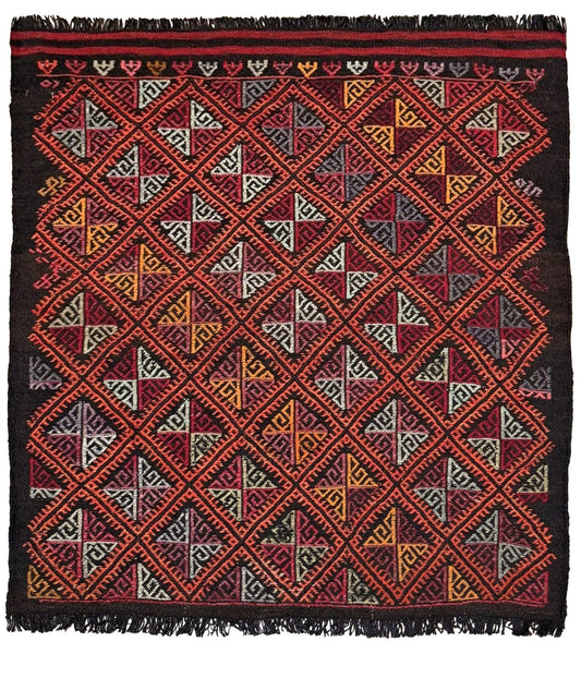 Decorative square turkish kilim rug with geometric pattern on a wooden floor, black chair, and potted plant in the background.