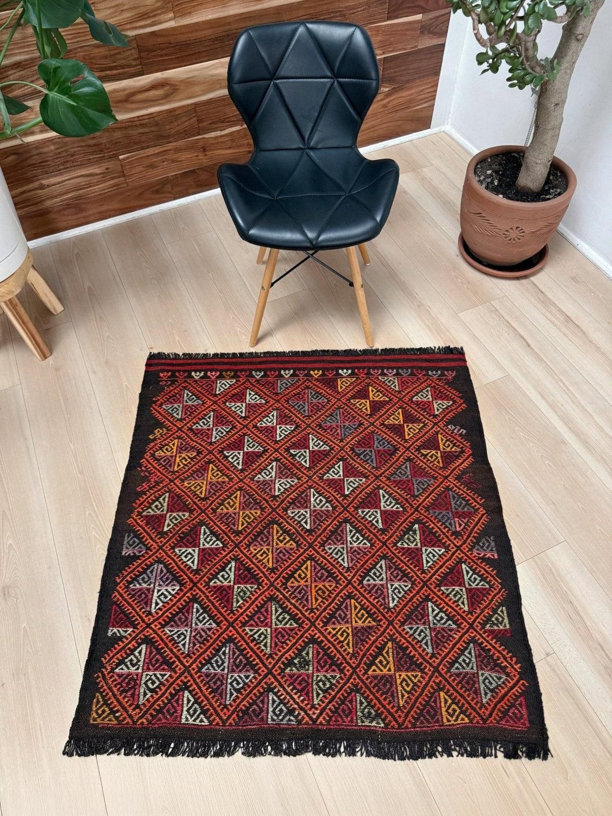 A square kilim area rug with a geometric diamond pattern in red, black, beige, and yellow, styled on a light wood floor in a room setting with a black chair and potted plants.