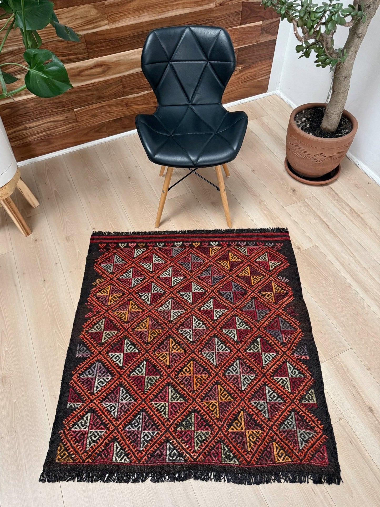 Decorative rug with geometric pattern on a wooden floor, black chair, and potted plant in the background.
