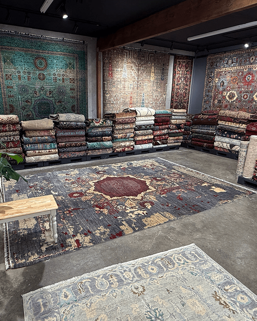 A large vintage-style area rug with a distressed navy, red, and beige pattern is displayed on a concrete floor in a showroom, with stacks of folded rugs and other wall-hung rugs visible in the background.