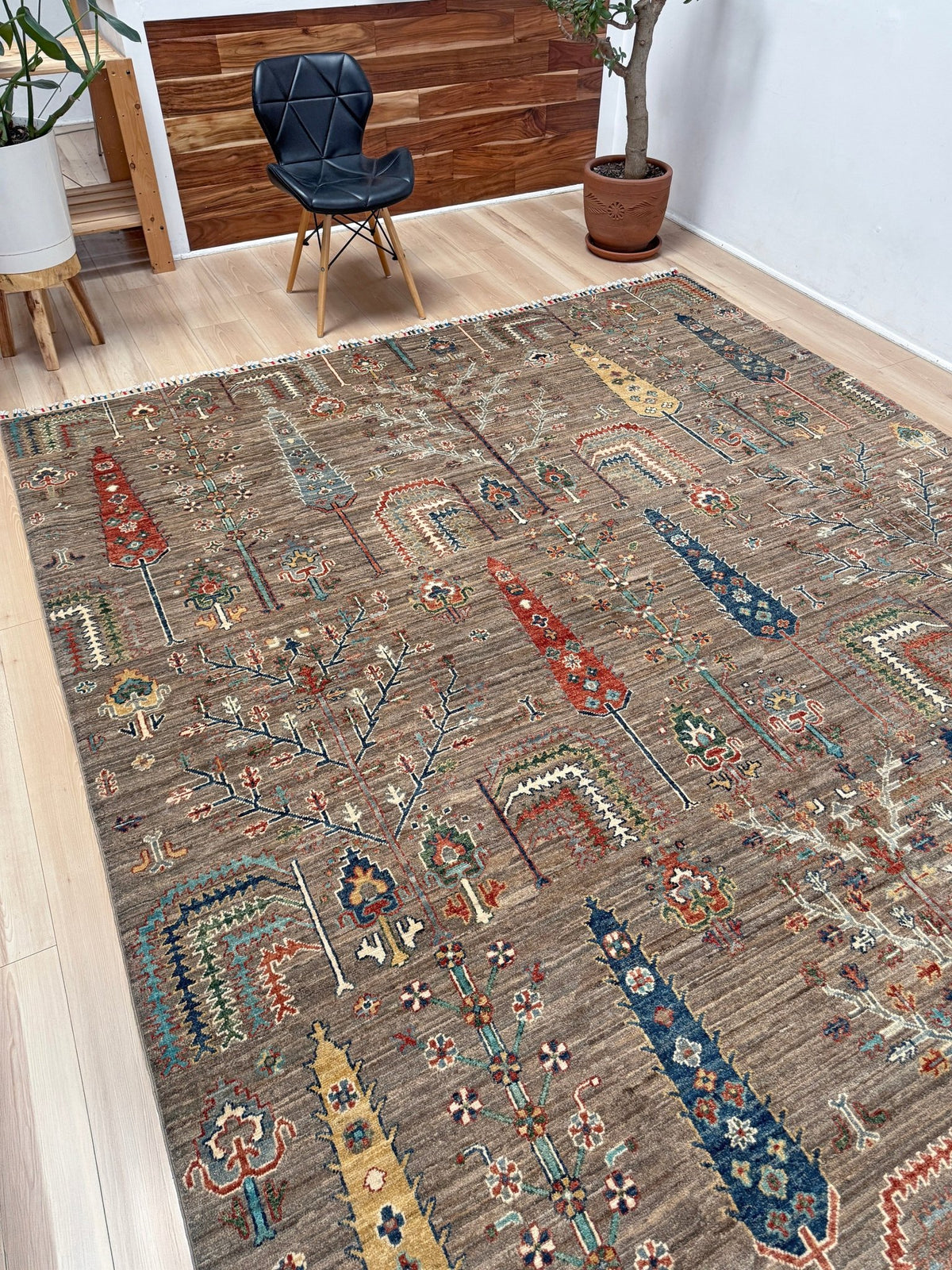 A large rectangular area rug with an intricate tribal-style pattern in brown, beige, blue, red, and yellow, shown in a modern living room setting with a black chair and potted plants.