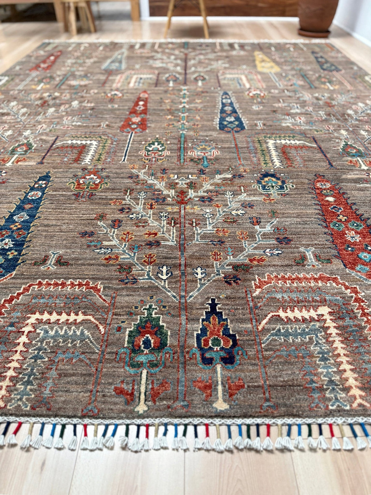 A traditional hand-knotted area rug with a complex geometric and floral pattern, displayed on a light-colored wooden floor. The rug features a dominant taupe-brown base with accents of red, blue, green, and cream. 8x10 Taupe Tree of Life Turkish rug with borderless floral garden design, stylized trees, handmade wool from Indigo Rugs Burlingame Bay Area.