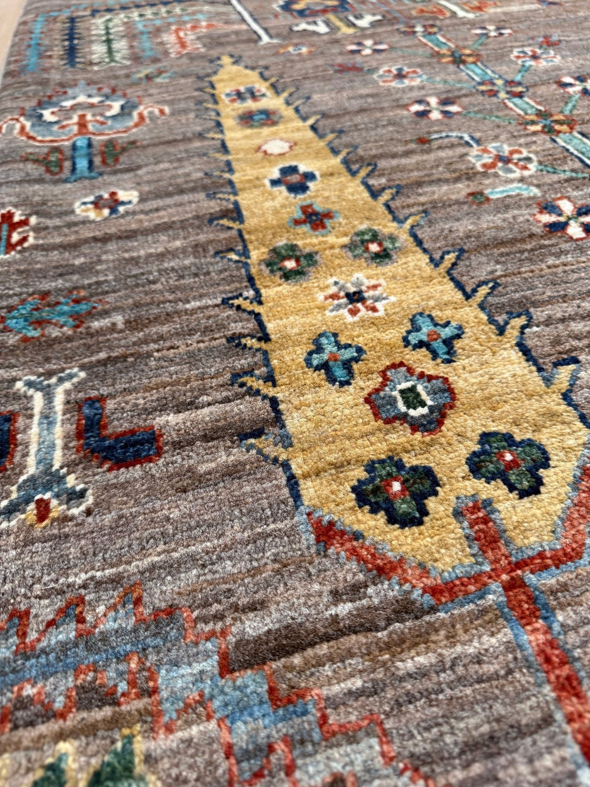 A close-up of a patterned area rug with a textured, low-pile surface. The design features a central, elongated pointed motif in a warm beige/tan color, decorated with blue, green, and red floral details, set against a mottled gray-brown background with additional blue and red motifs.