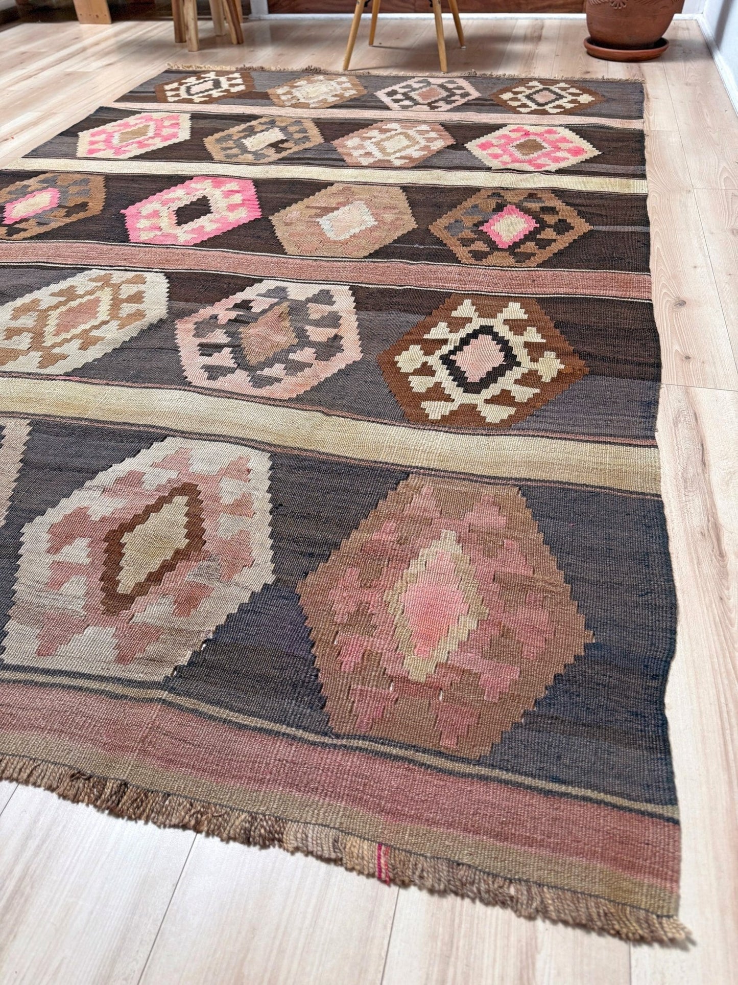 A rectangular woven area rug with a Southwestern geometric pattern, laid out on a light wood floor. The rug features a dark charcoal gray base with large pink, beige, and brown diamond motifs and fringe along the bottom edge. Handmade wool 6x9 rug from Indigo Rugs Burlingame Bay Area.