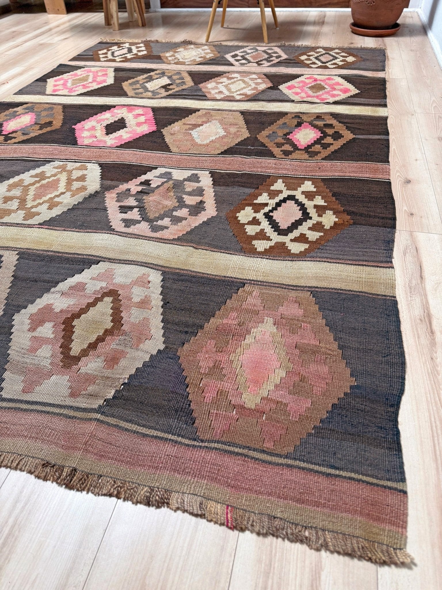 A rectangular woven area rug with a Southwestern geometric pattern, laid out on a light wood floor. The rug features a dark charcoal gray base with large pink, beige, and brown diamond motifs and fringe along the bottom edge. Handmade wool 6x9 rug from Indigo Rugs Burlingame Bay Area.