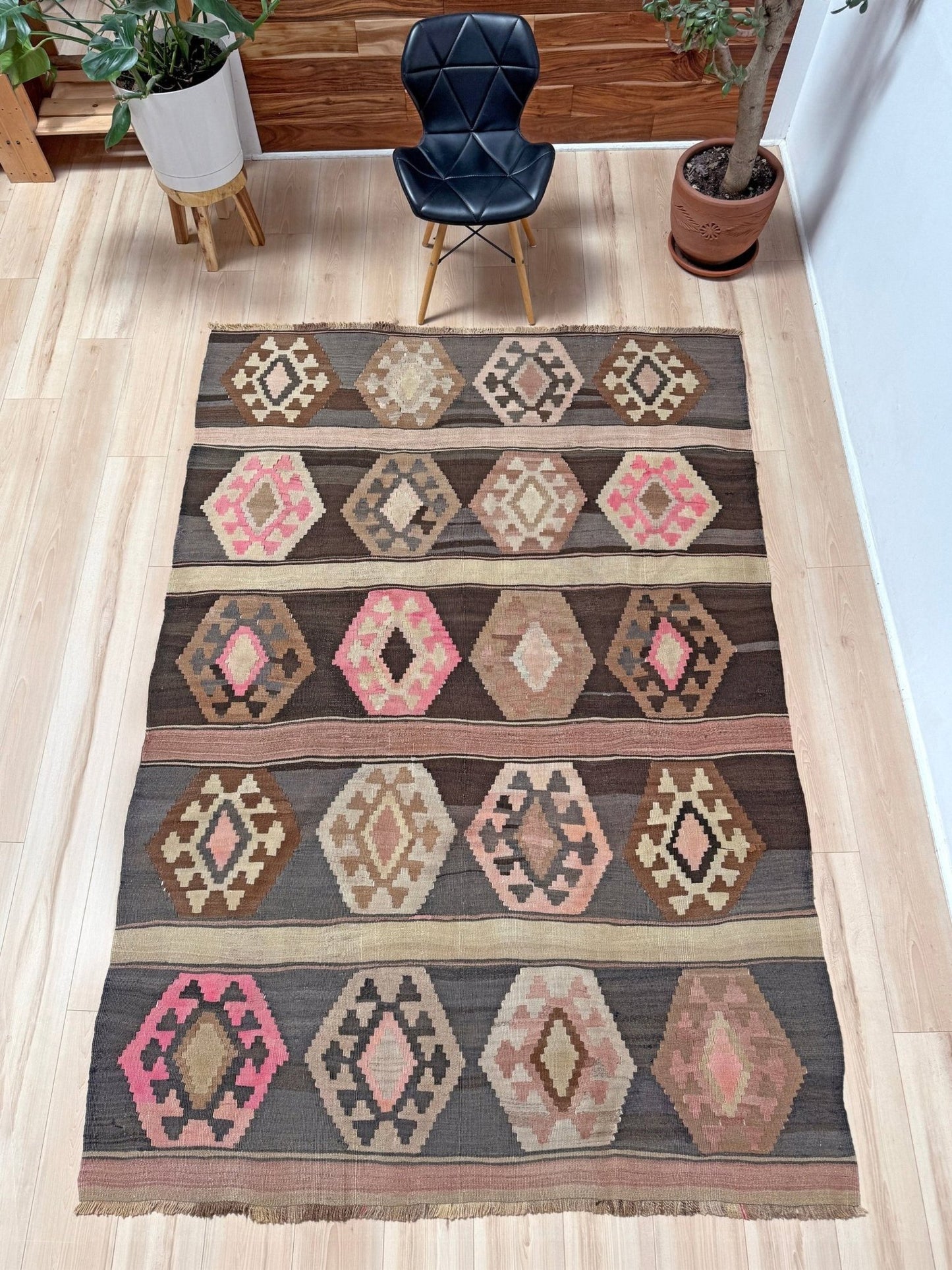 A rectangular woven area rug with a geometric tribal pattern in brown, beige, pink, and gray, displayed on a light wood floor in a room setting with a black chair and potted plants. Handmade wool 6x9 rug from Indigo Rugs Burlingame Bay Area.