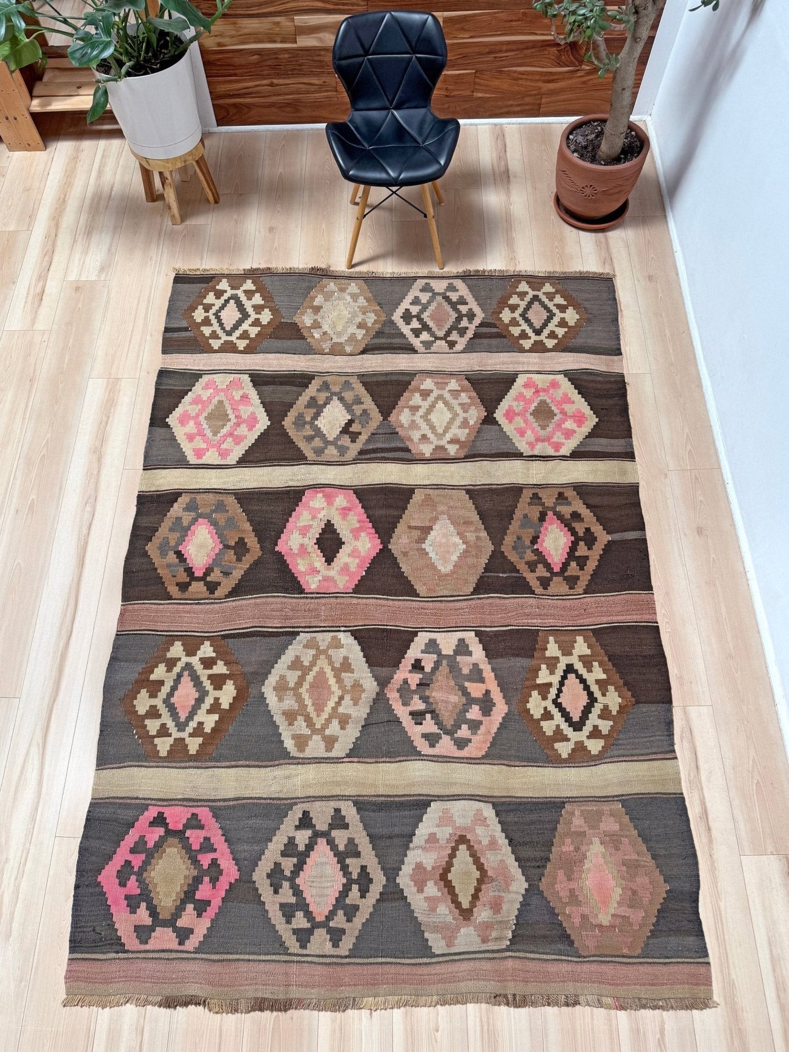 A rectangular woven area rug with a geometric tribal pattern in brown, beige, pink, and gray, displayed on a light wood floor in a room setting with a black chair and potted plants. Handmade wool 6x9 rug from Indigo Rugs Burlingame Bay Area.