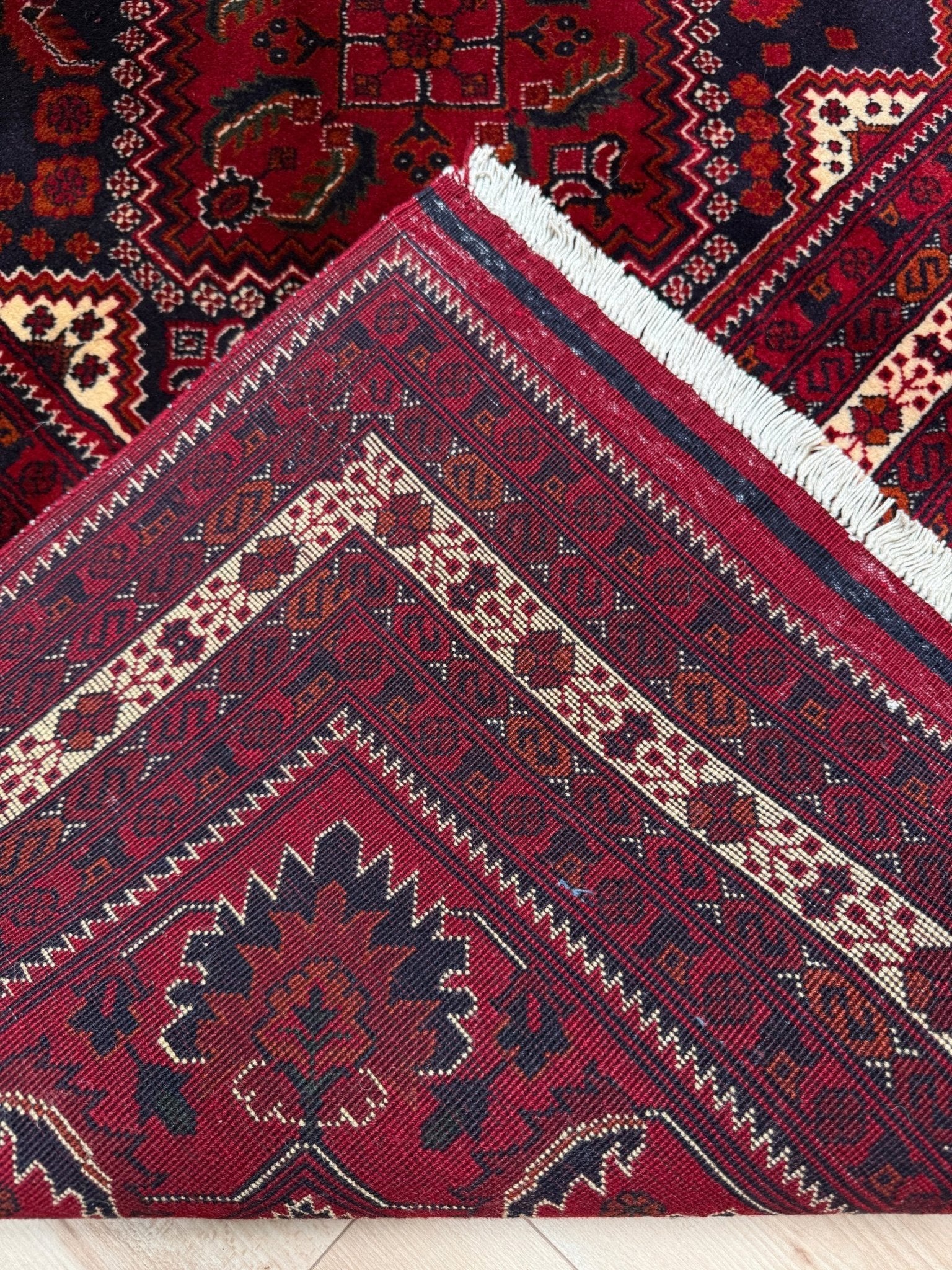 A close-up of a traditional patterned rug corner showing a deep red field with cream, black, and brown geometric borders, a white fringe edge, and part of another patterned rug underneath.