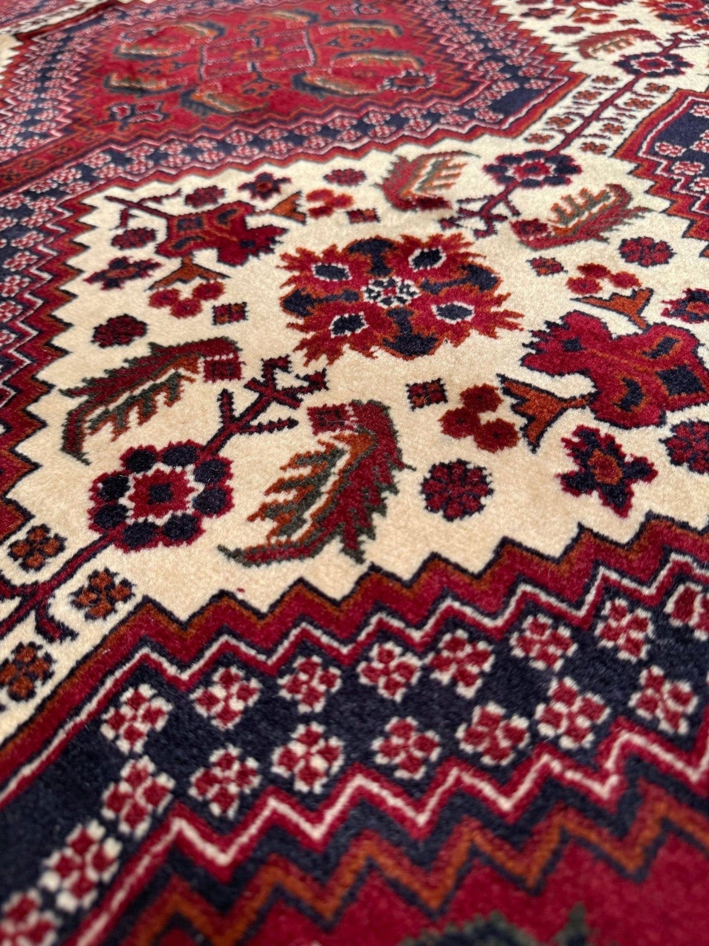 A close-up view of a traditional patterned rug with a cream central field, red and navy floral motifs, and geometric borders in red, navy, and cream.