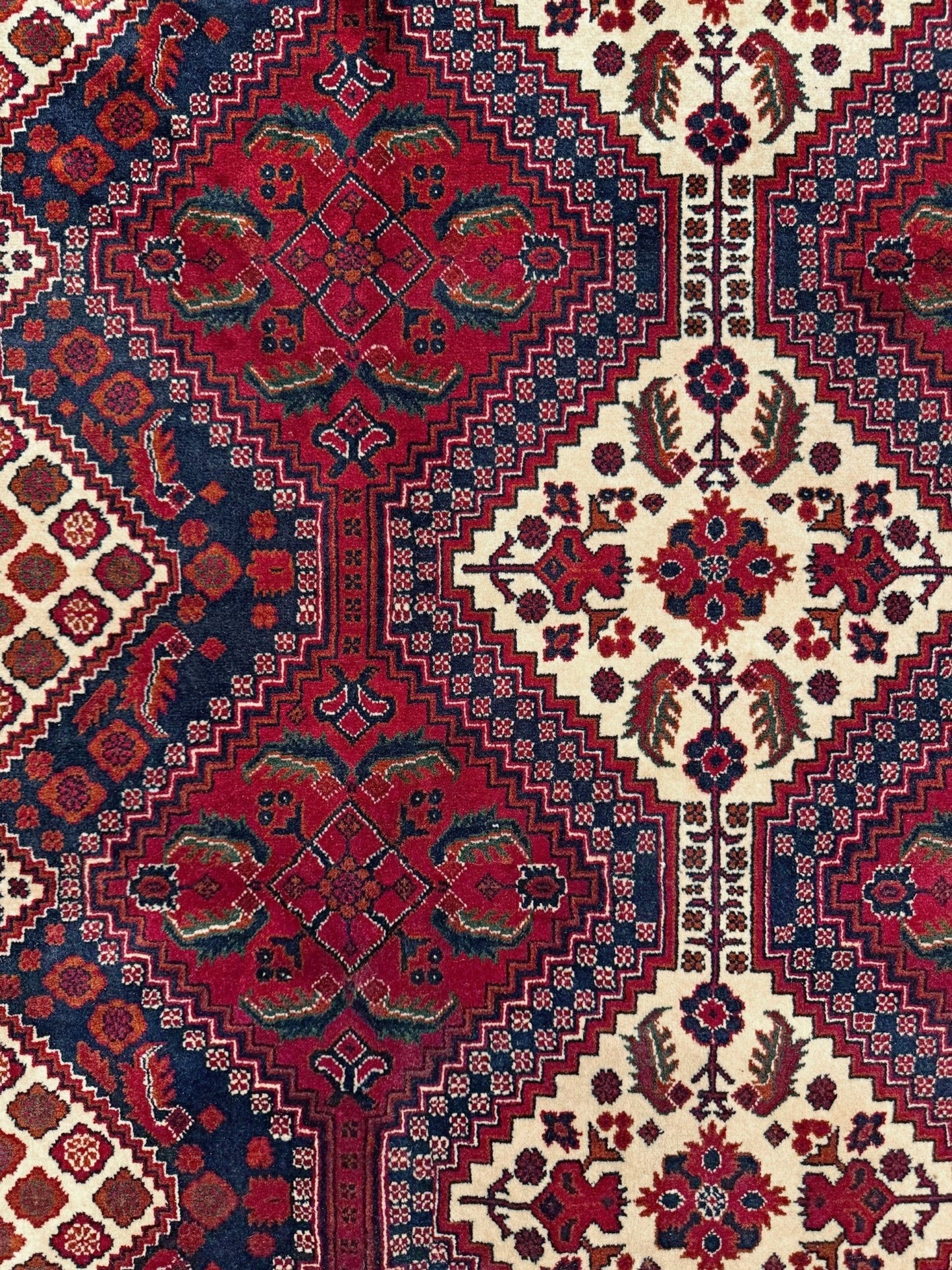 A close-up of a traditional patterned rug with a red central field, cream side panels, and dark blue borders featuring geometric and floral motifs.