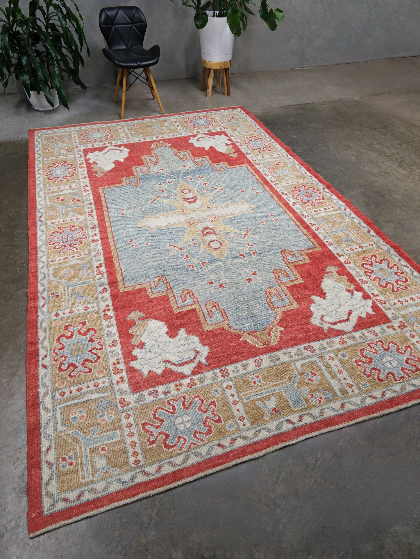 A rectangular area rug with a traditional geometric pattern, featuring a light blue center, red borders, and beige and cream motifs, displayed on a concrete floor in a room setting.