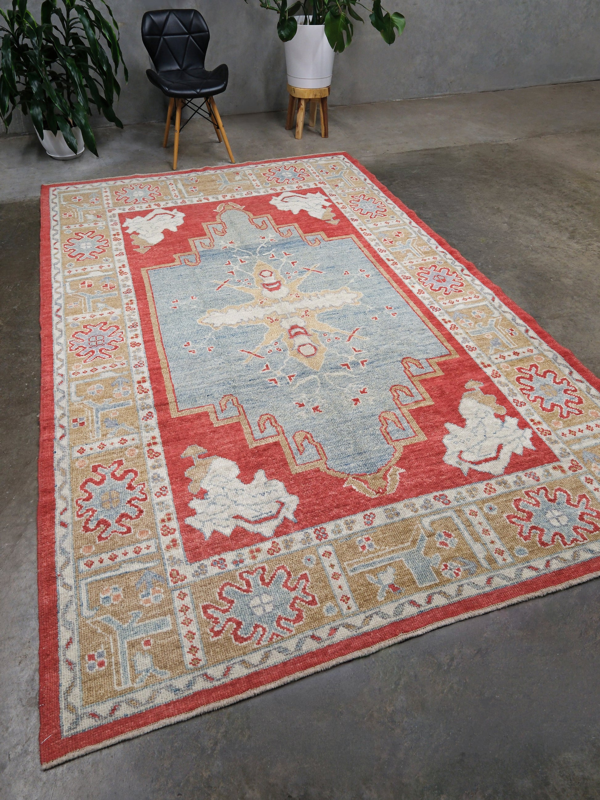 A rectangular area rug with a traditional geometric pattern, featuring a light blue center, red borders, and beige and cream motifs, displayed on a concrete floor in a room setting.