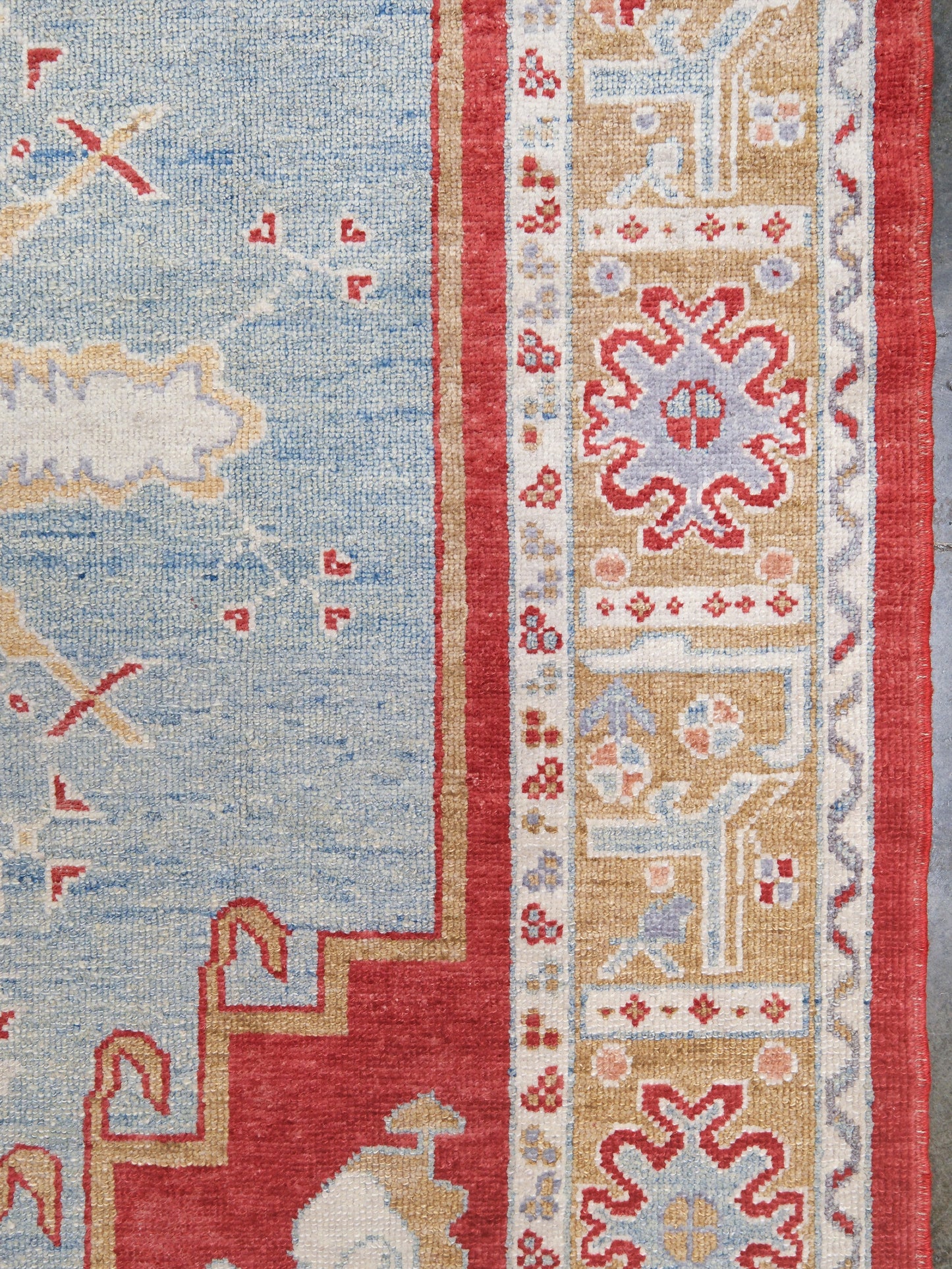 Close-up of a patterned rug showing a light blue field, red border details, and a beige central panel with cream and red motifs.