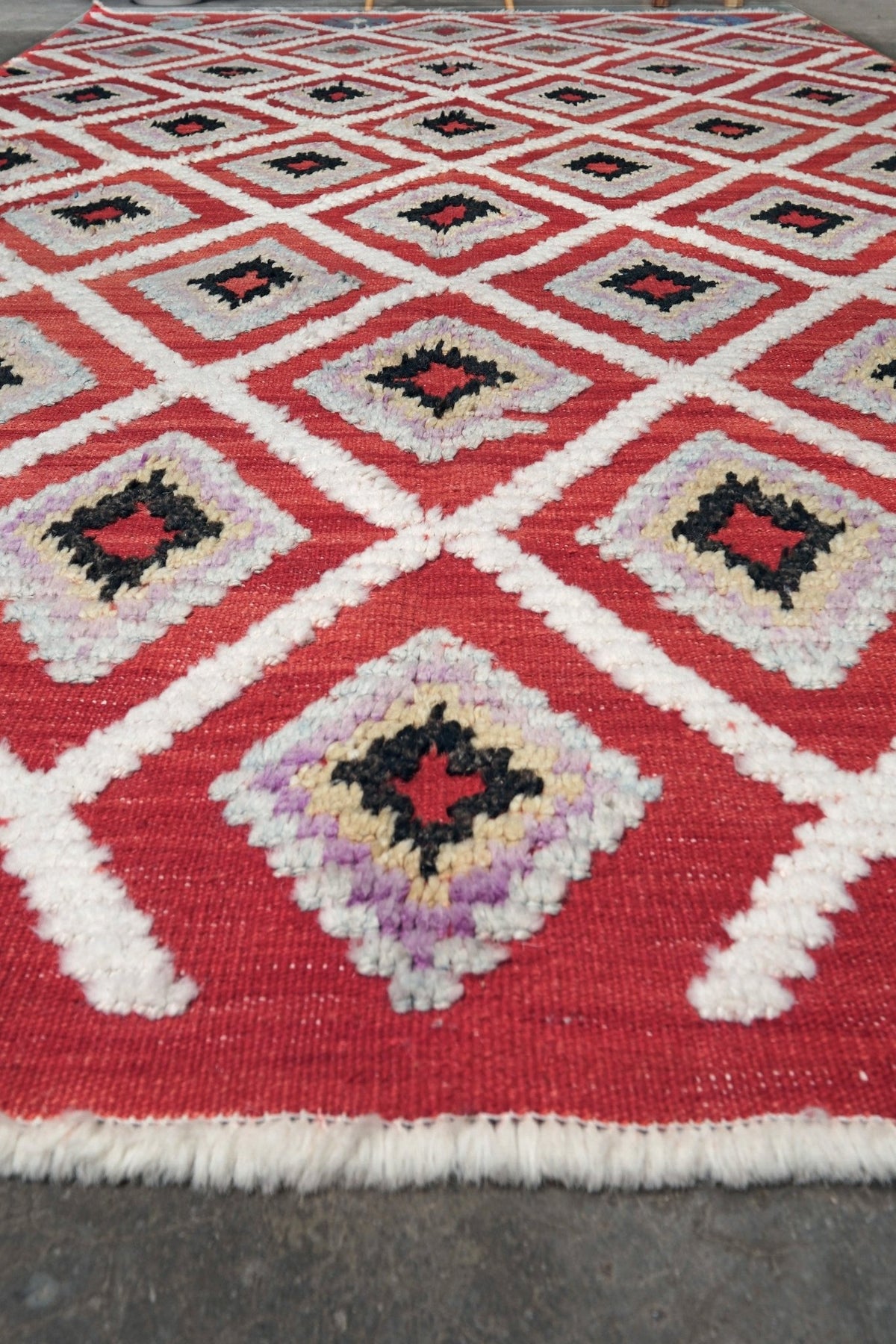 A close-up of a woven geometric area rug with a red field, white diamond borders, and central motifs in black, gray, purple, and cream, laid flat on a dark surface.