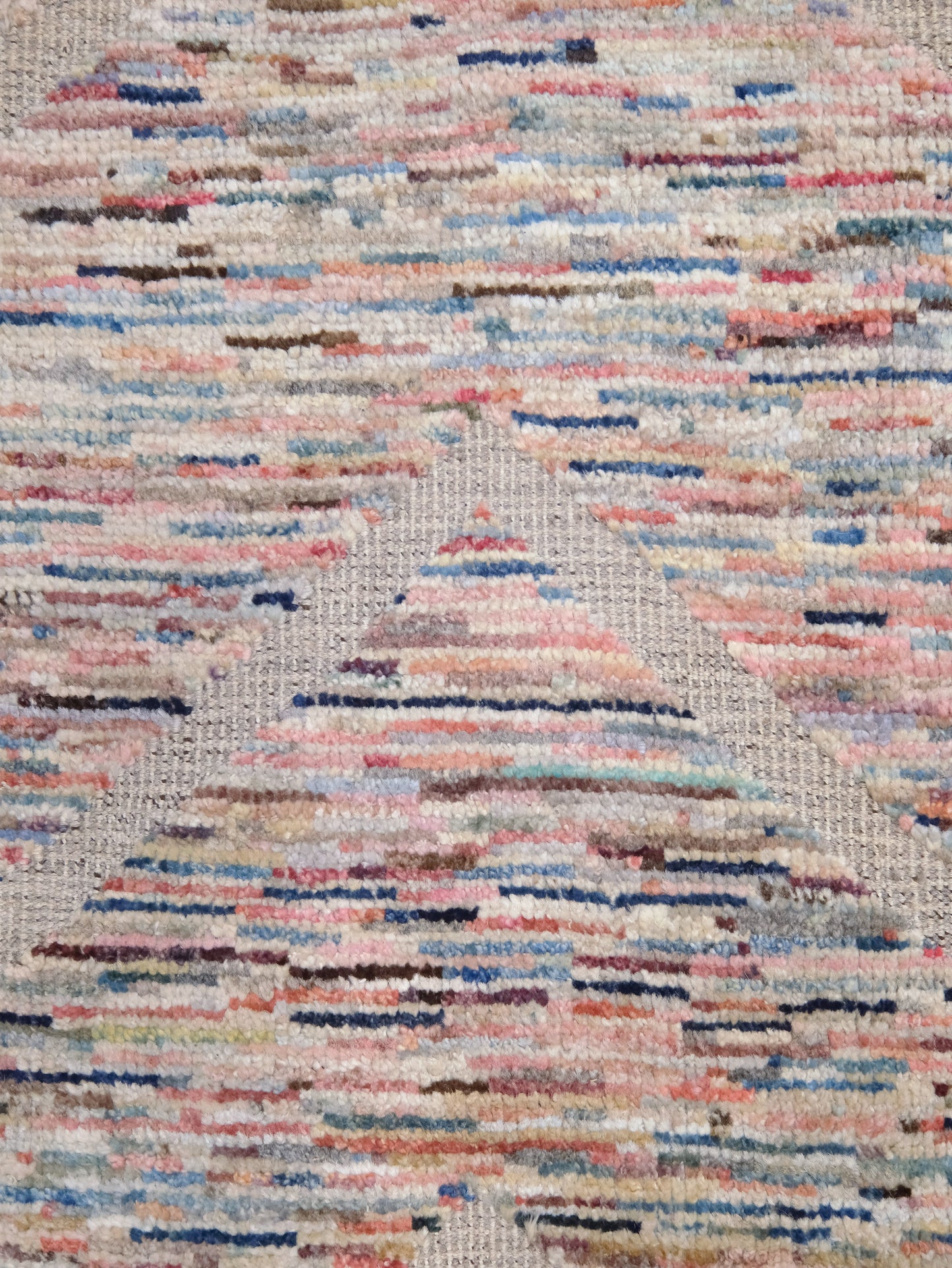 A close-up of a textured woven rug with a multicolor abstract pattern of horizontal stripes in pink, blue, beige, and brown on a light gray base.