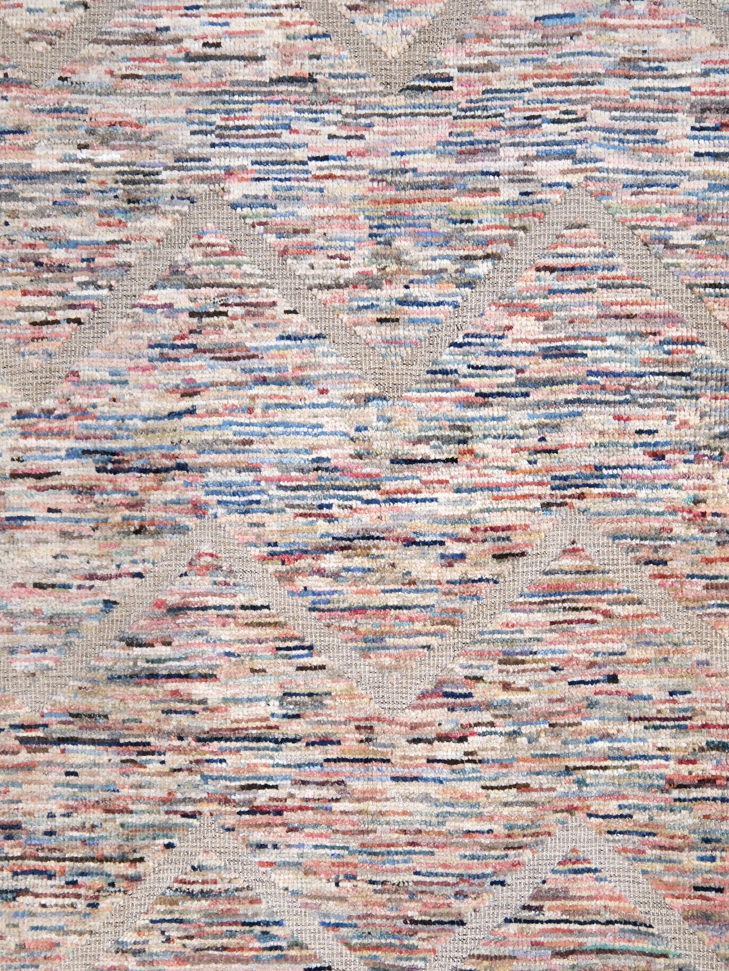 A close-up of a textured woven rug or fabric with a multicolor abstract pattern of pink, blue, beige, and brown horizontal lines over a light beige background.