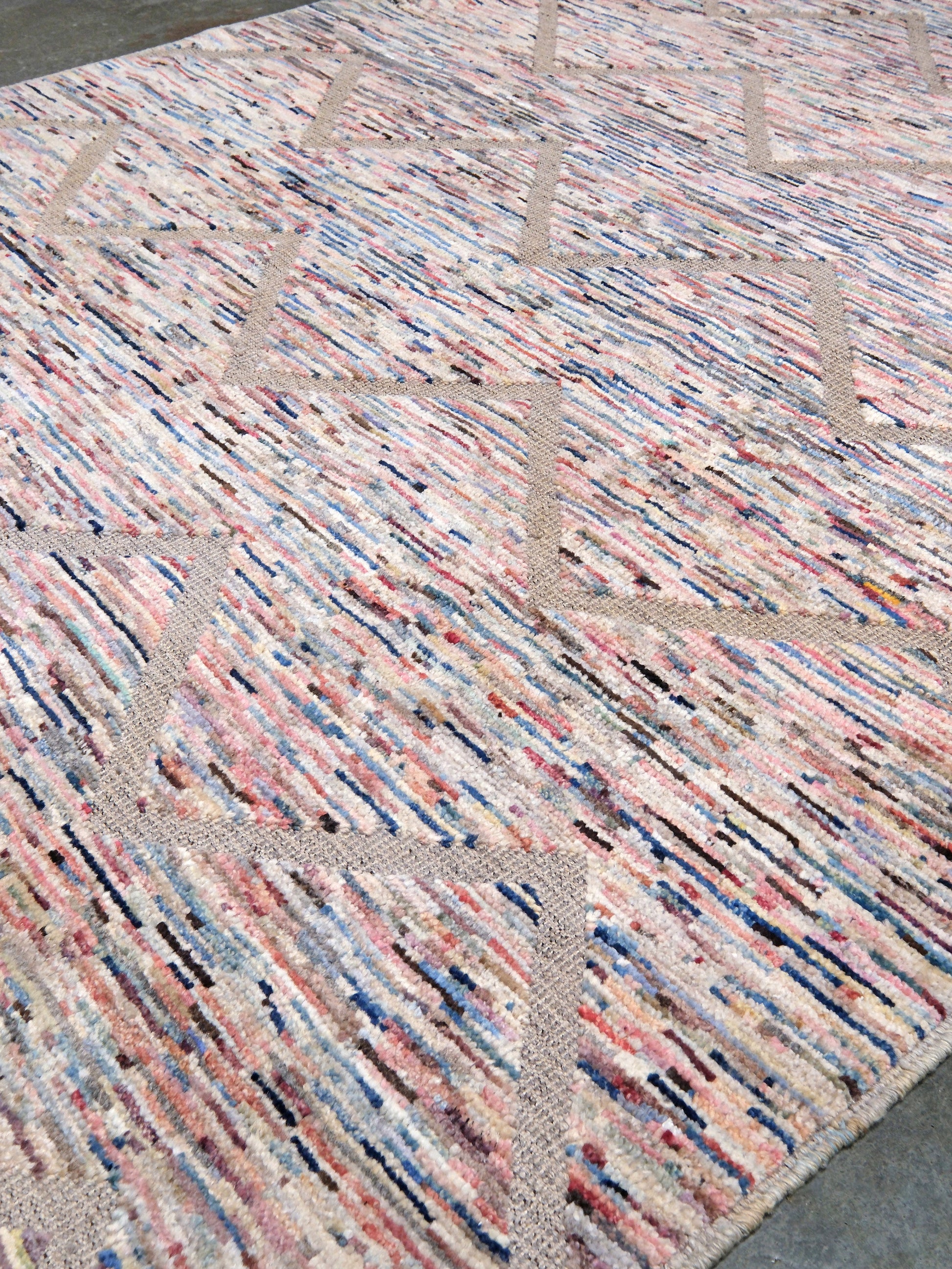 A close-up of a textured area rug with a multicolor abstract pattern of pink, blue, beige, and black lines on a cream base, laid out on a gray concrete floor.