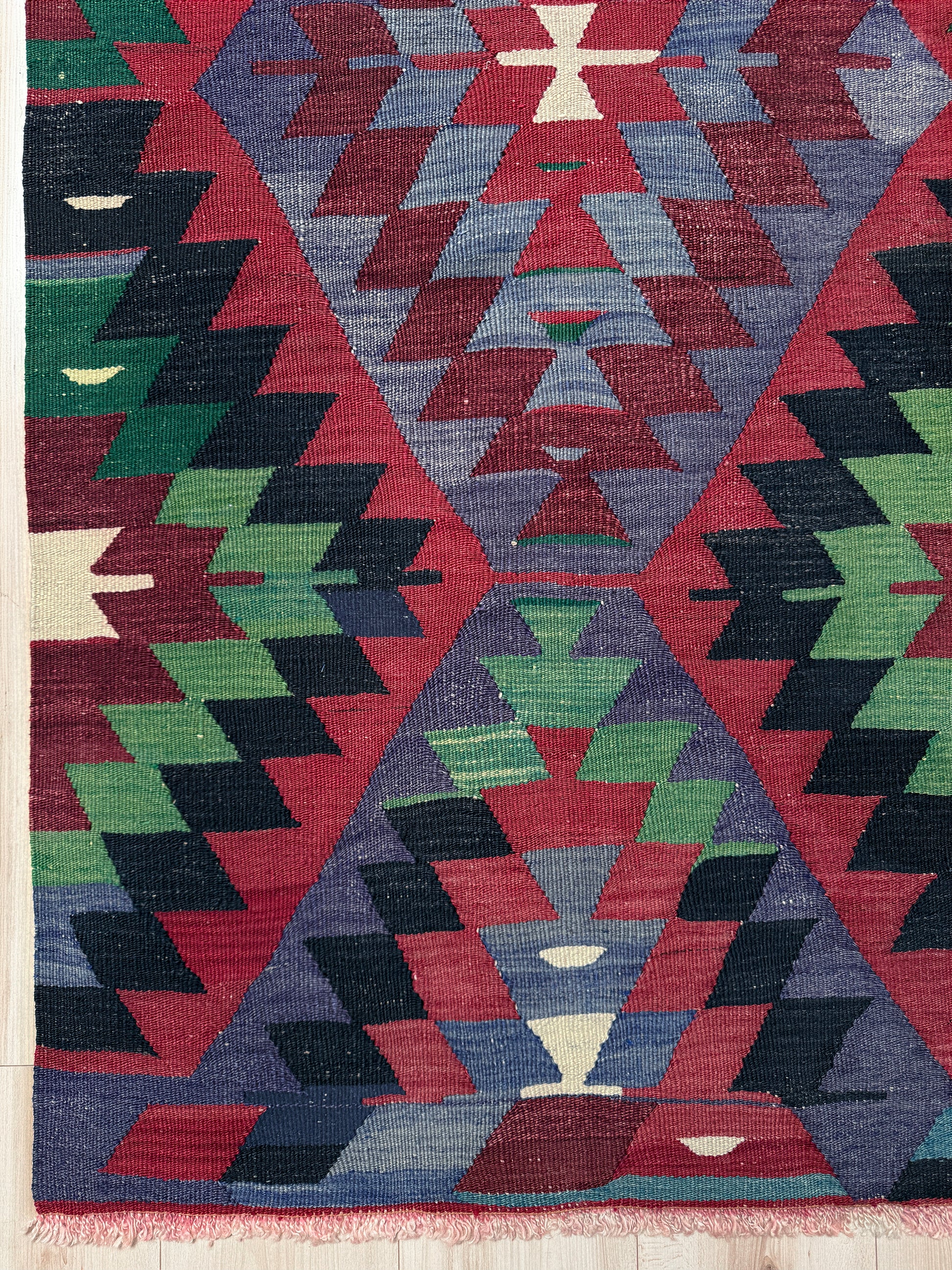 Geometric patterned rug with triangles in red, blue, green, and black on a wooden floor. Vintage Esme Turkish tribal kilim rug featuring geometric design in raspberry red, blue violet, emerald green, handmade 6x10 perfect for boho style 