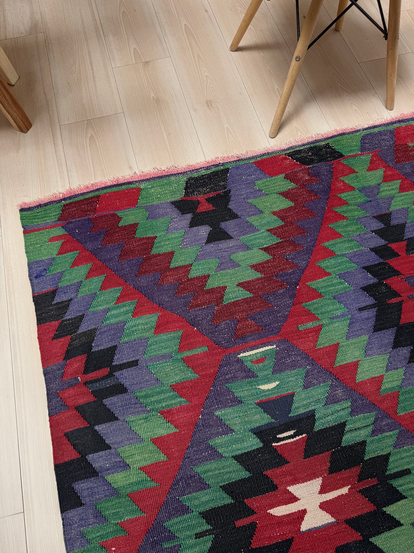 Colorful geometric-patterned rug on a light wooden floor with a chair partially visible. Vintage Esme Turkish tribal kilim rug featuring geometric design in raspberry red, blue violet, emerald green, handmade 6x10 perfect for boho style 