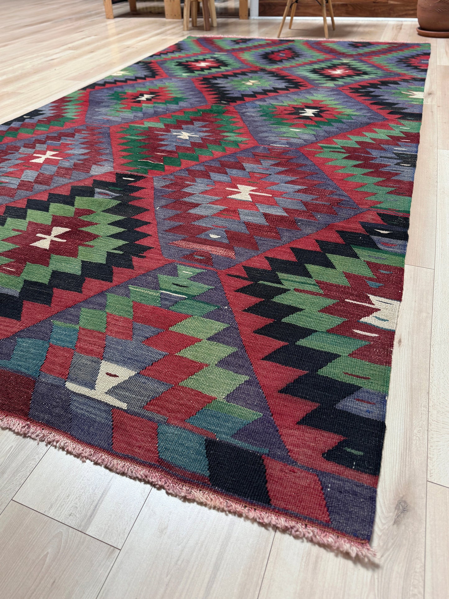 Multicolored geometric patterned rug on a wooden floor. Vintage Esme Turkish tribal kilim rug featuring geometric design in raspberry red, blue violet, emerald green, handmade 6x10 perfect for boho style 