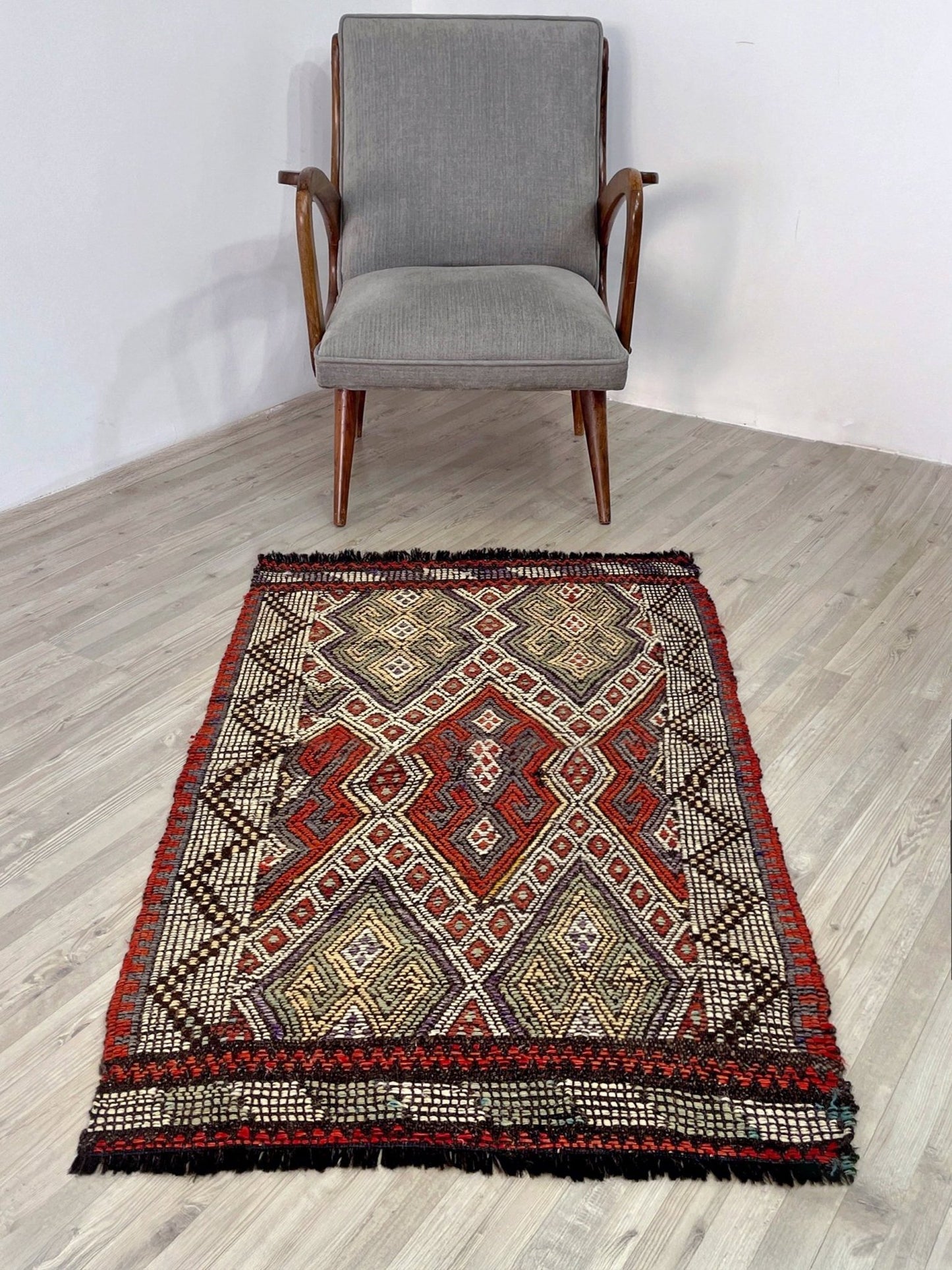 Decorative rug with geometric pattern on a wooden floor, chair in the background. Small vintage Turkish cicim kilim rug, 2x4, with raised tribal embroidery in rust red, ivory, olive, and charcoal on a goat hair foundation.