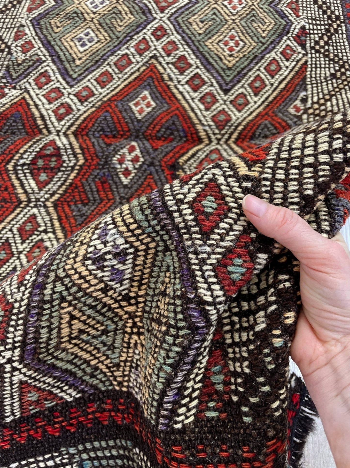 Close-up of a hand holding patterned fabric with geometric designs. Small vintage Turkish cicim kilim rug, 2x4, with raised tribal embroidery in rust red, ivory, olive, and charcoal on a goat hair foundation.