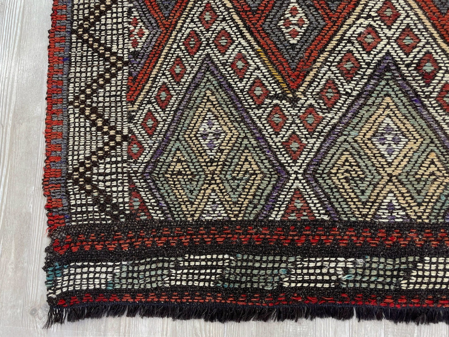 Close-up of a patterned rug with geometric designs on a wooden floor. Small vintage Turkish cicim kilim rug, 2x4, with raised tribal embroidery in rust red, ivory, olive, and charcoal on a goat hair foundation.